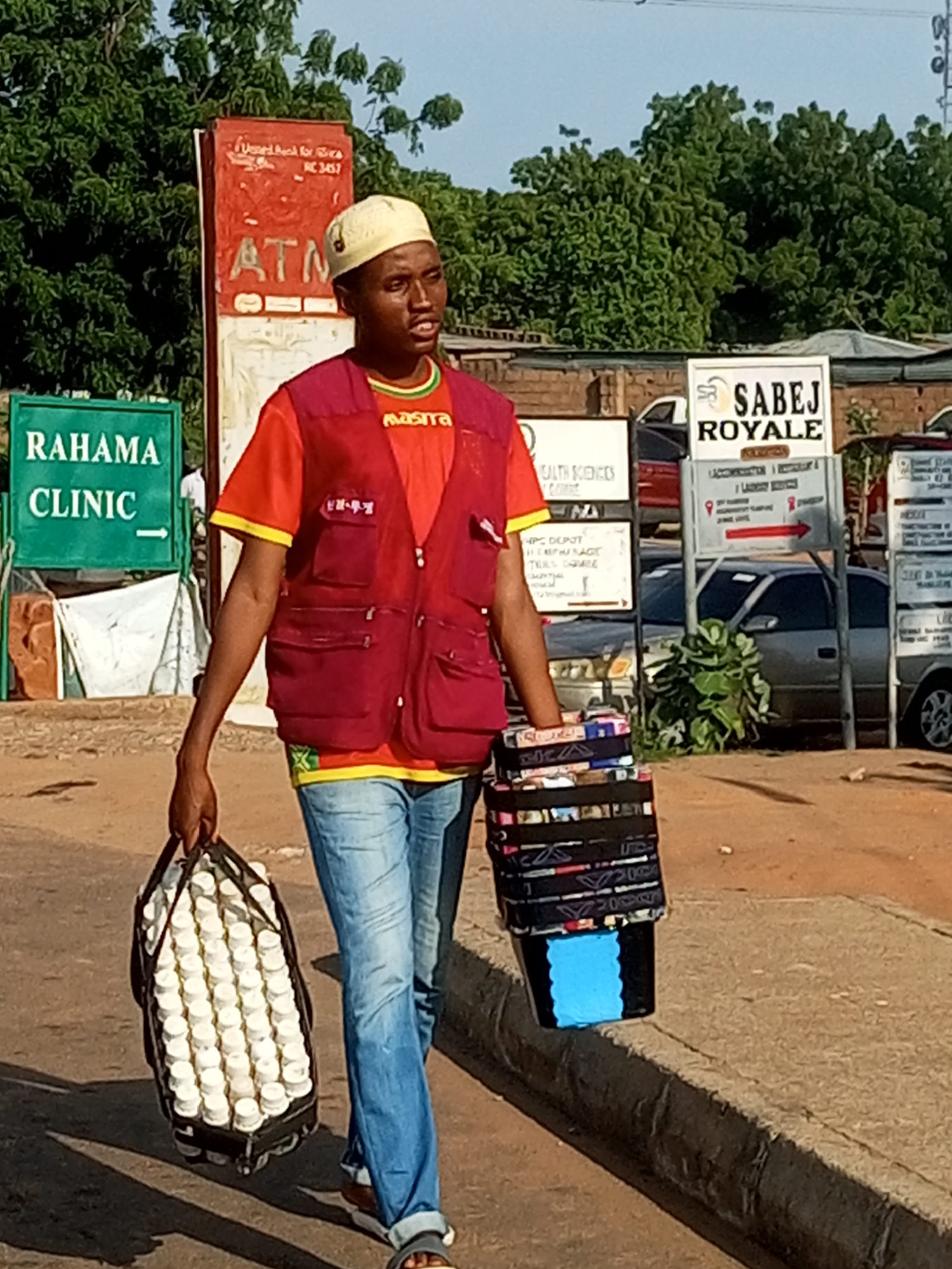 This media file captures a man walking down a street in Gombe, Nigeria, carrying two items. He is wearing a red vest over an orange shirt and blue jeans, with a light-colored cap on his head. In his right hand, he carries a black carrier filled with many small, white-capped bottles, likely for sale. In his left hand, he holds another black carrier containing what appear to be books or boxes.
In the background, signs provide context for the location. One green sign reads "RAHAMA CLINIC" with an arrow pointing to the right. Another sign features "SABEJ ROYALE" in large letters. An ATM machine is also visible on a red structure. The scene appears to be outdoors, under a bright sky, suggesting it is daytime. The trees and brick buildings in the background are consistent with an urban or semi-urban environment in Nigeria. The man's expression is neutral as he focuses on his task of carrying his goods.