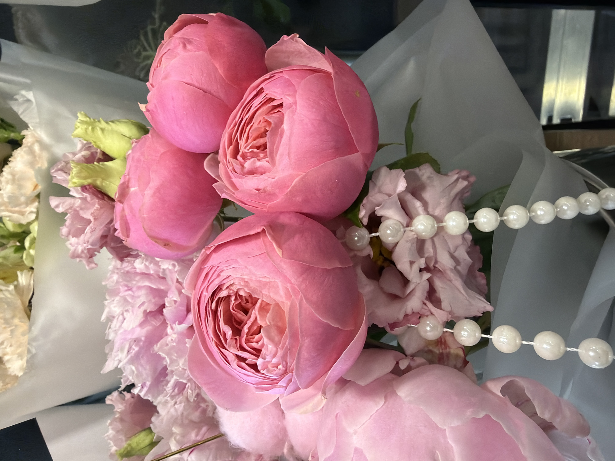 This media file features a close-up shot of a bouquet of pink roses and other flowers. The roses are large and fully bloomed, with many delicate petals. There are also some lighter pink, frilly flowers and green buds visible. A strand of pearls is draped across the bouquet, adding a touch of elegance. The flowers are wrapped in light-colored paper, possibly tissue paper or cellophane, with a subtle sheen. The background is blurred and dark, with some indistinct shapes that might be part of a shelf or display. The lighting is soft and appears to be artificial, creating a warm and inviting atmosphere. The focus is entirely on the floral arrangement, highlighting the textures and colors of the blooms. There is no visible text in the image. The overall impression is one of natural beauty and a sophisticated aesthetic, possibly intended for a special occasion or as a decorative element.