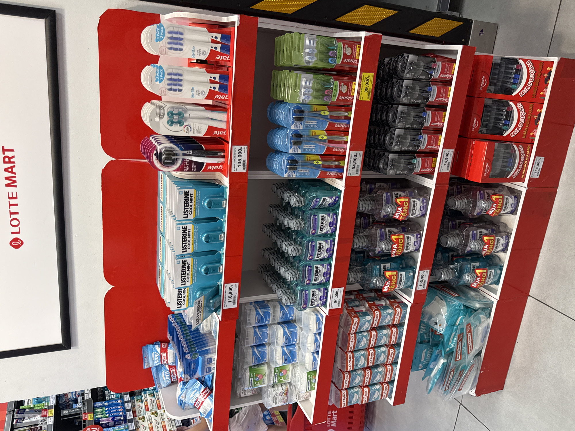 The image shows a retail display of oral hygiene products at a Lotte Mart in Nha Trang, Vietnam. The display features multiple shelves filled with various brands and types of toothbrushes, mouthwash, and other dental care items. prominent brands include Listerine and Colgate. Some products have price tags displayed in Vietnamese Dong. The overall environment appears to be a well-lit supermarket aisle. There are no people clearly visible in the frame.