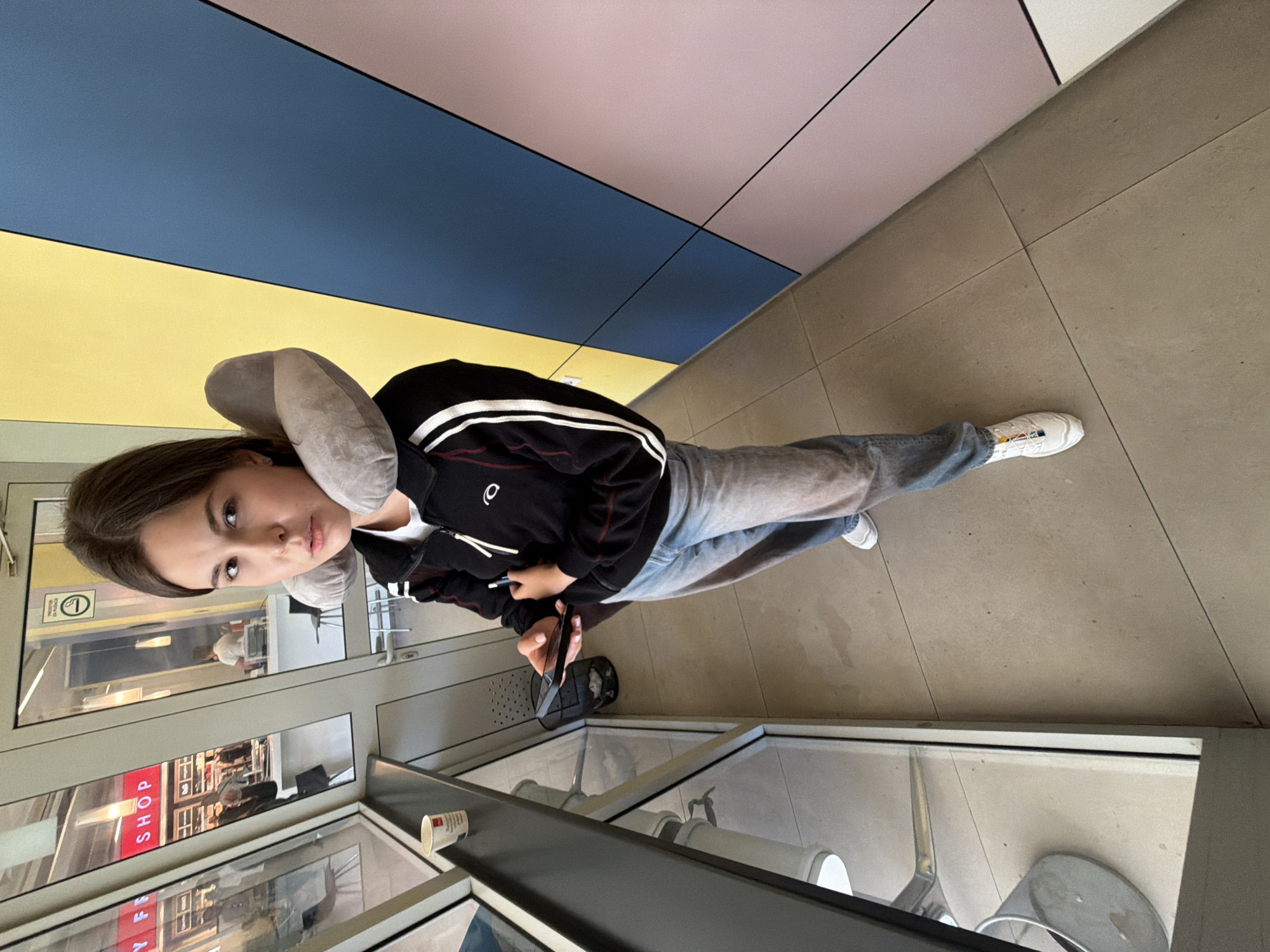 A young woman wearing a black jacket with white stripes, light blue jeans, and white sneakers stands indoors next to a window, holding a mobile phone. She has a gray travel pillow around her neck. Reflected in the window are shops and signs with the word "SHOP" visible. A small black bag is near her feet. The floor is tiled, and the background includes a wall or ceiling divided into sections of yellow, blue, and pink. The setting appears to be a waiting area in a commercial building. There is no information to indicate this image was taken in Gornje Dubrave, Bosnia and Herzegovina.