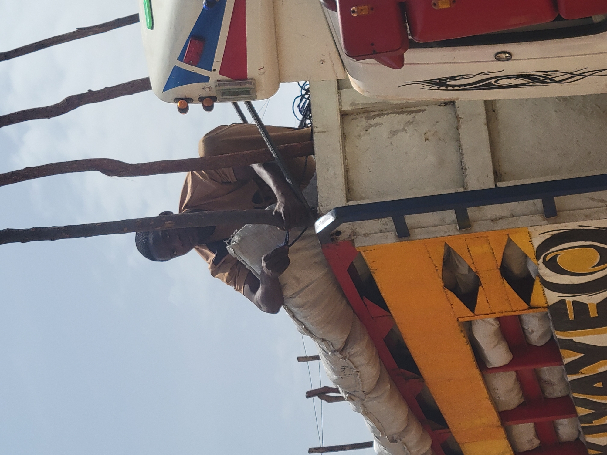 A man is perched on a wooden structure, possibly involved in construction or repair work. He is wearing a tan shirt and light-colored pants. The structure has elements of scaffolding, constructed with raw wood. The environment includes parts of a carnival or amusement ride, with sections painted in orange, red, white, and black, featuring stylized designs. The sky is visible in the background. Based on the context cues, the location is likely Jos, Nigeria.