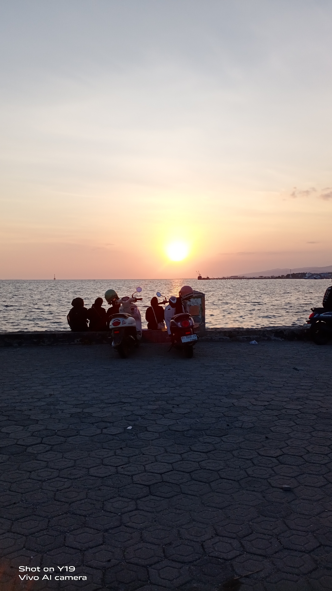 The image depicts a serene sunset scene at the waterfront in the city of Sea, Indonesia. Several individuals are seated on a low wall overlooking the ocean, accompanied by three scooters parked nearby. The sun, a bright orb, hangs low in the sky, casting a warm, golden glow across the water and the horizon. The sky transitions from a soft orange near the sun to a pale blue at the top of the frame. In the distance, landmasses with distant structures and a sailboat are visible. The foreground consists of a paved area with a hexagonal pattern. The overall atmosphere is peaceful and tranquil, capturing a moment of shared appreciation for the natural beauty of the seascape.