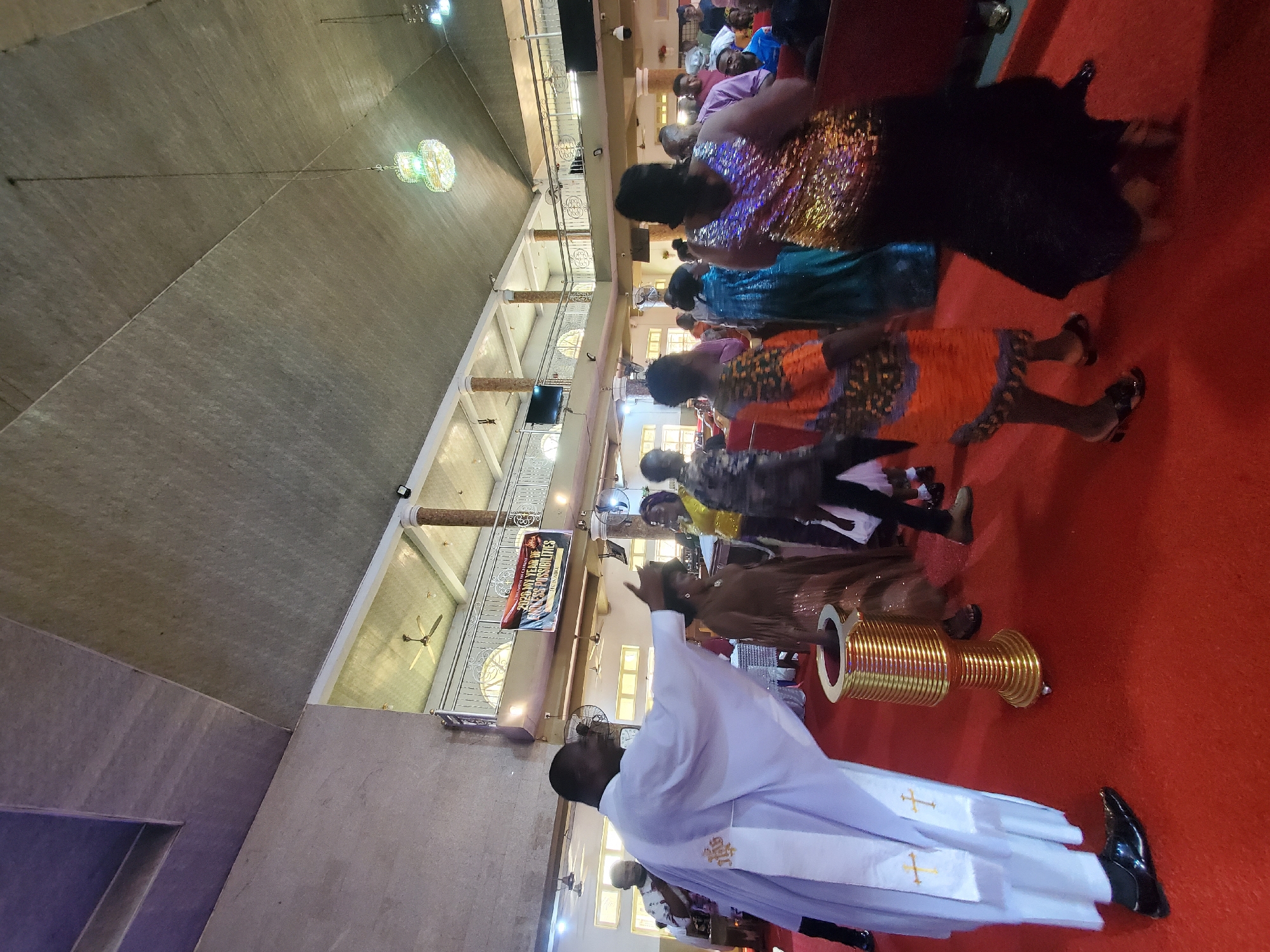 A male dressed in a white robe with crosses on the sash is conducting a church service in Onitsha, Nigeria. Attendees are in colorful clothes, lined up in pews. A large golden chalice sits on a red carpet near the man. Chandeliers hang from the ceiling.