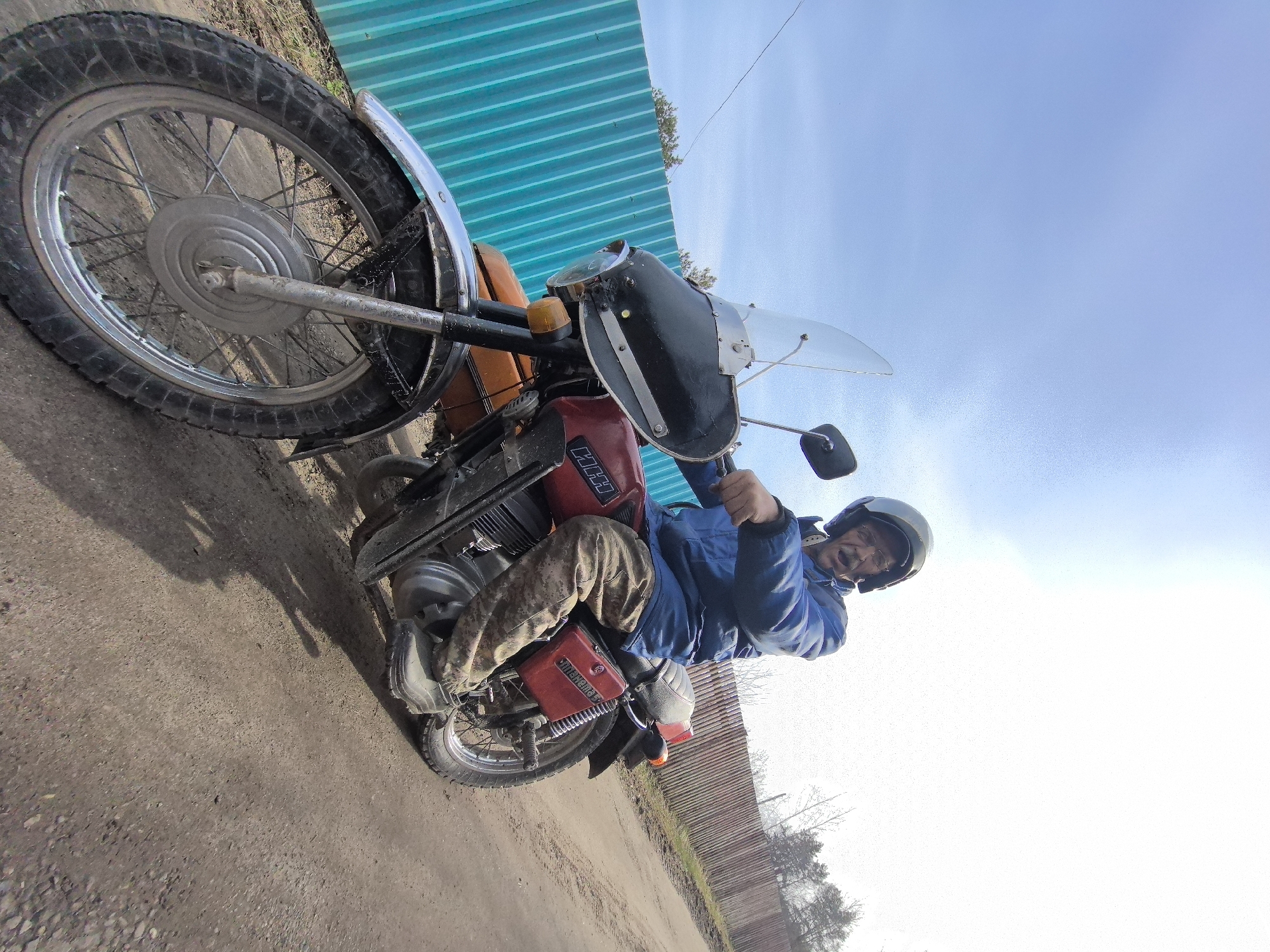 A man is sitting on a vintage motorcycle. The motorcycle is red and black with a windshield. The man is wearing a blue jacket, camouflage pants, and a helmet. The ground is dirt, and there is a blue fence in the background. The sky is partly cloudy. The location appears to be rural or semi-rural, possibly on the outskirts of Noviy, Russia, given the presence of the weathered fence and dirt ground.
