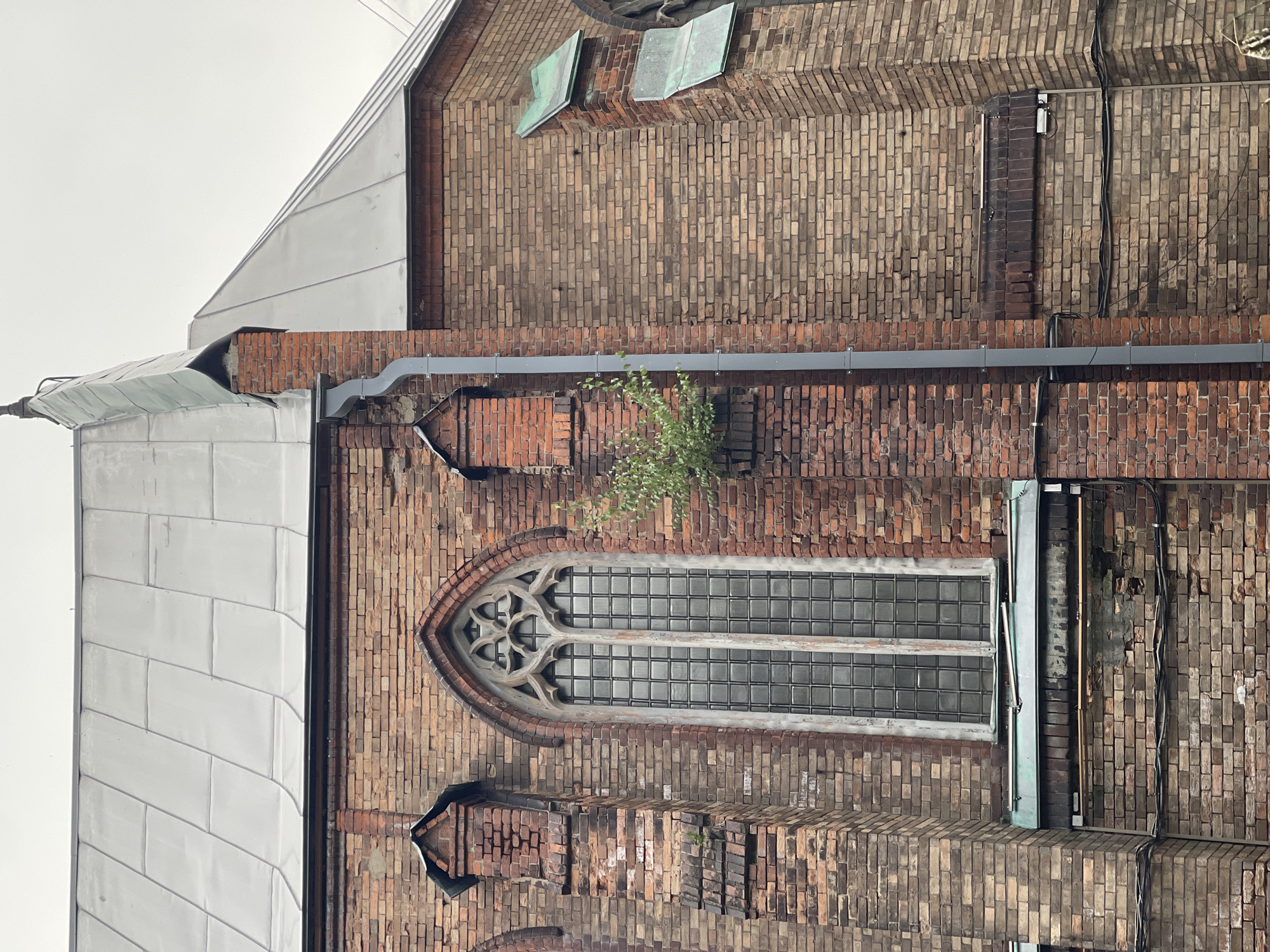 The image shows the exterior of a brick building with a large, arched window. The building is constructed from weathered bricks of varying shades of red and brown. The window has a Gothic-style stone tracery at the top and is filled with small, square glass panes. A thin tree with green leaves grows out of the brickwork near the window.
To the left of the brick wall, there is a section of a white, angular roof made of metal panels. Above the brick wall, two square, weathered copper-colored skylights or vents are visible. The overall impression is of an old building, possibly a church or a historic structure, showing signs of age and exposure to the elements. The lighting is diffused, suggesting an overcast day. There are no people visible in the image. The scene appears to be a close-up, focusing on architectural details and the texture of the materials. The orientation of the image is vertical.