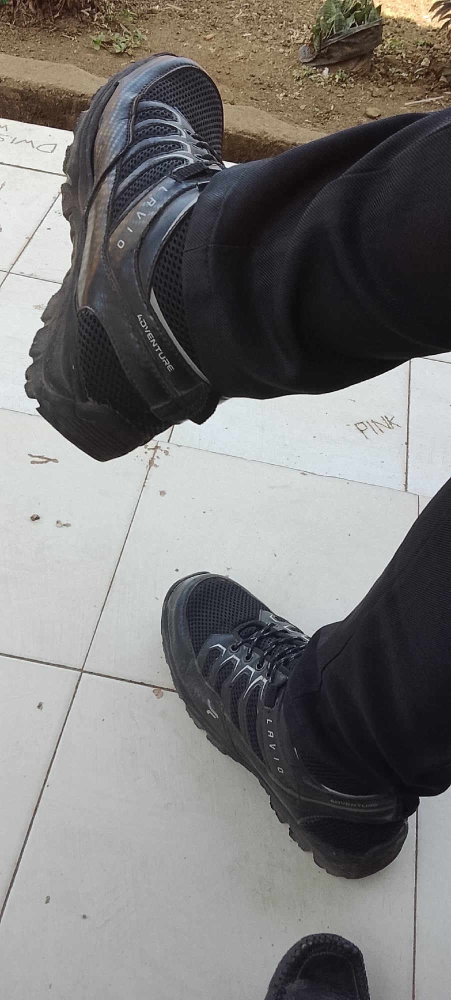 In Tanjung Setia, Indonesia, a person is sitting down, wearing black pants and black athletic shoes. The shoes have "LAVIO" and "ADVENTURE" written on them. The scene appears to be outdoors, on a tiled surface, with dirt and plants visible in the background. There are also some faint markings on the tiles, including the word "PINK".