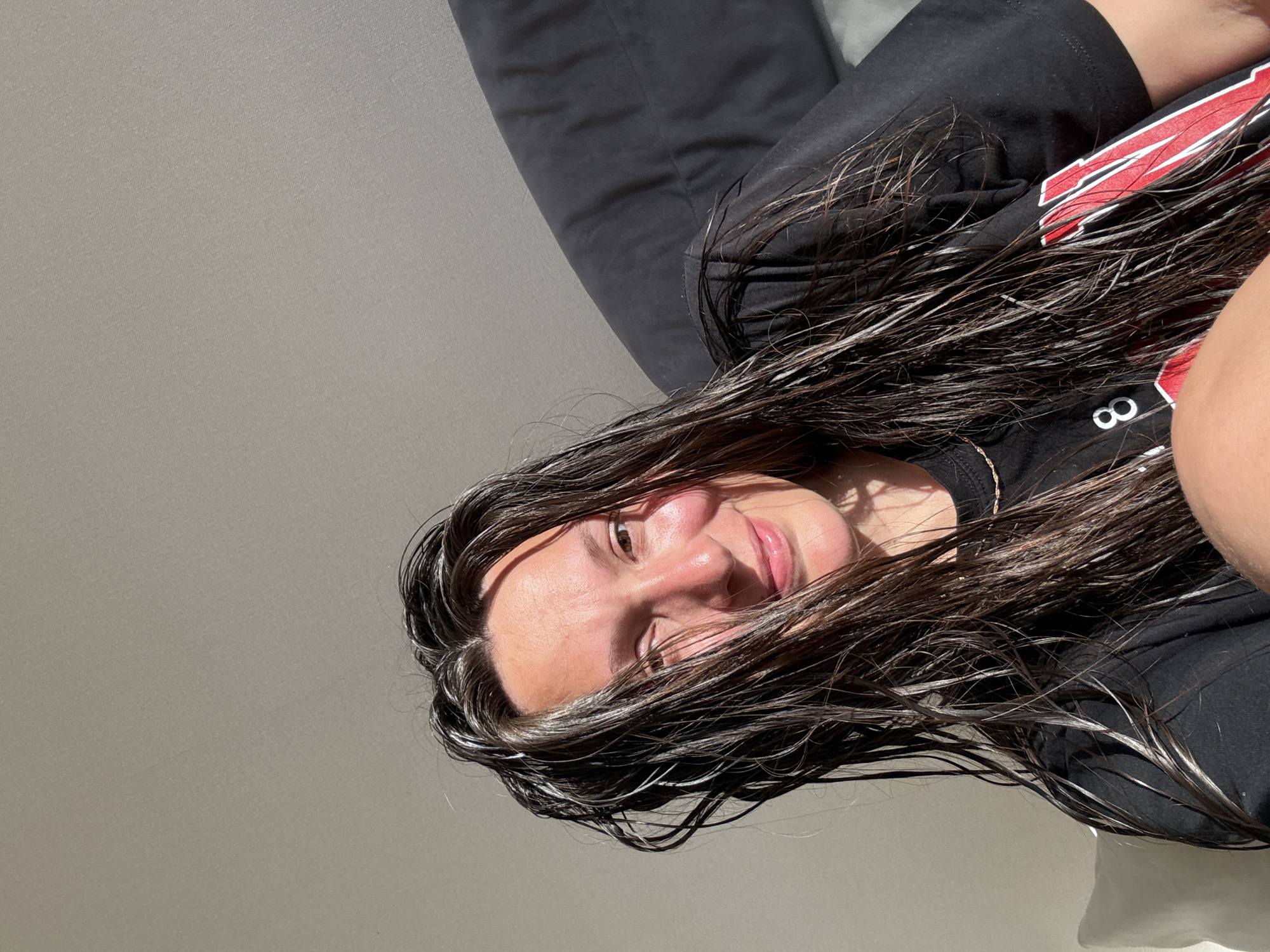 This close-up portrait features a woman with long, dark, wet hair, smiling softly at the camera. Her hair, parted on her left, cascades around her face and shoulders, appearing damp and exhibiting prominent highlights and some lighter strands, possibly from sun or natural color variations. She is wearing a black short-sleeved t-shirt with a red and white graphic design that includes partial numbers or letters, such as '8' or 'B'. A thin, light-colored necklace is visible around her neck. Strong natural light, likely sunlight, illuminates her face and hair from the upper left, casting highlights on her skin and creating shadows on the right side of her face. The background consists of a plain, light taupe-colored wall with a subtle texture and, to the right, a dark grey or black pillow or cushion, suggesting an indoor, domestic setting. The scene is located in a room, in the city of Chaikovsky, Russia.