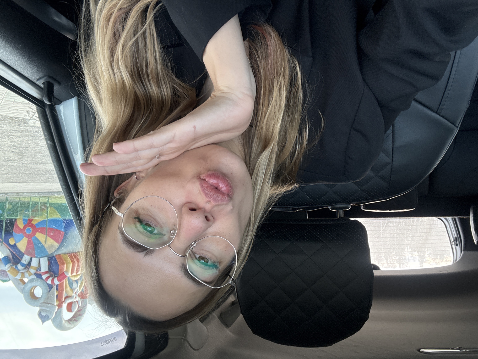 A woman with blonde hair, wearing glasses and a black jacket, is sitting in a car. She has her hand near her face and appears to be blowing a kiss. The car interior is visible, including a quilted leather headrest. Outside the car window, a colorful, decorative structure is visible. Based on the image details, the location is Ust-Ilimsk, Russia.