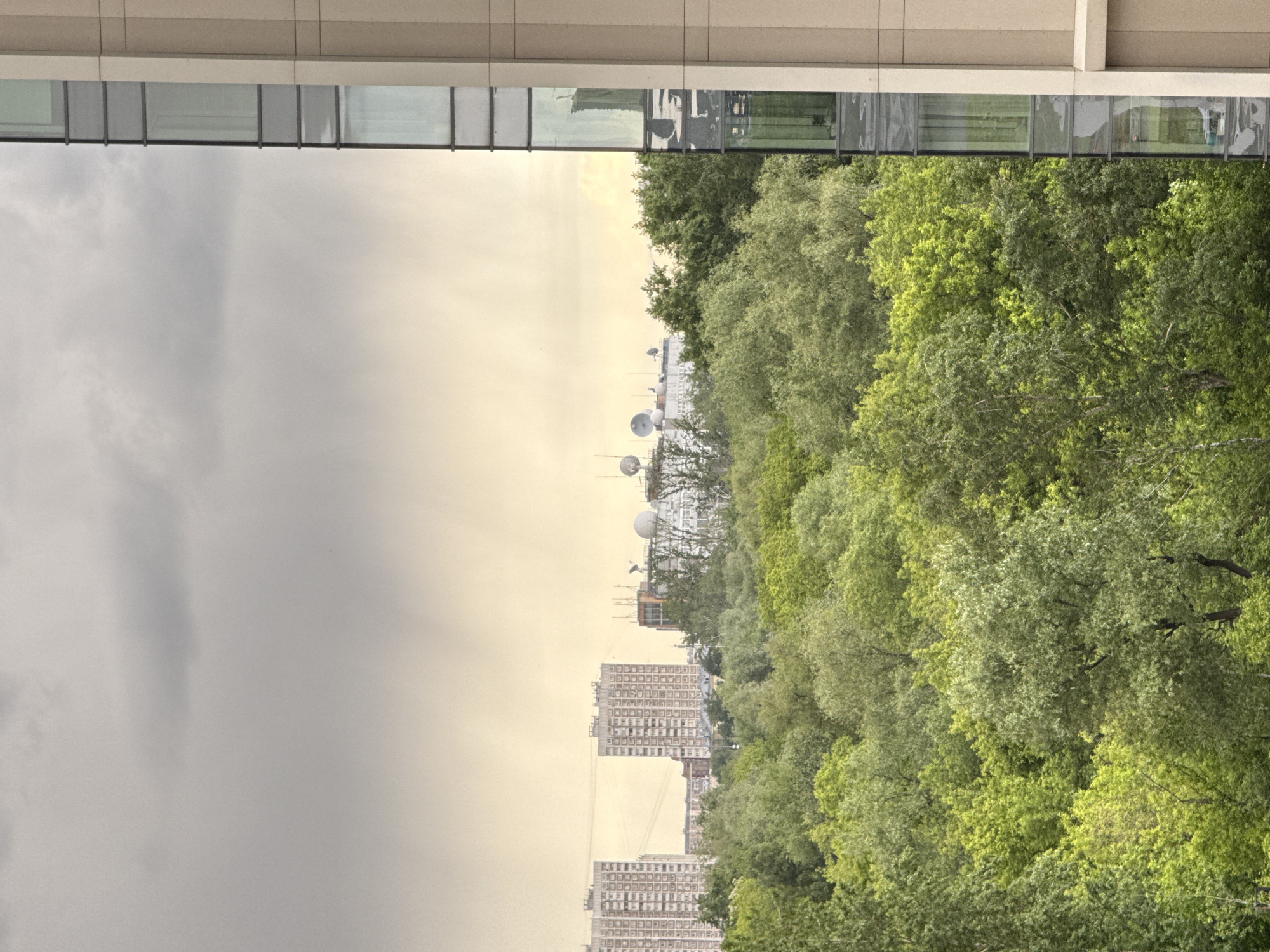 The image depicts a view from a high vantage point in Khimki, Russia. A section of a building with glass windows is visible at the top of the frame. Below, the scene transitions to a landscape featuring lush, green trees. In the distance, buildings including apartment blocks and a complex with spherical structures resembling radar domes are visible. The sky on the left is overcast and grey, contrasting with a lighter, yellowish hue towards the horizon.