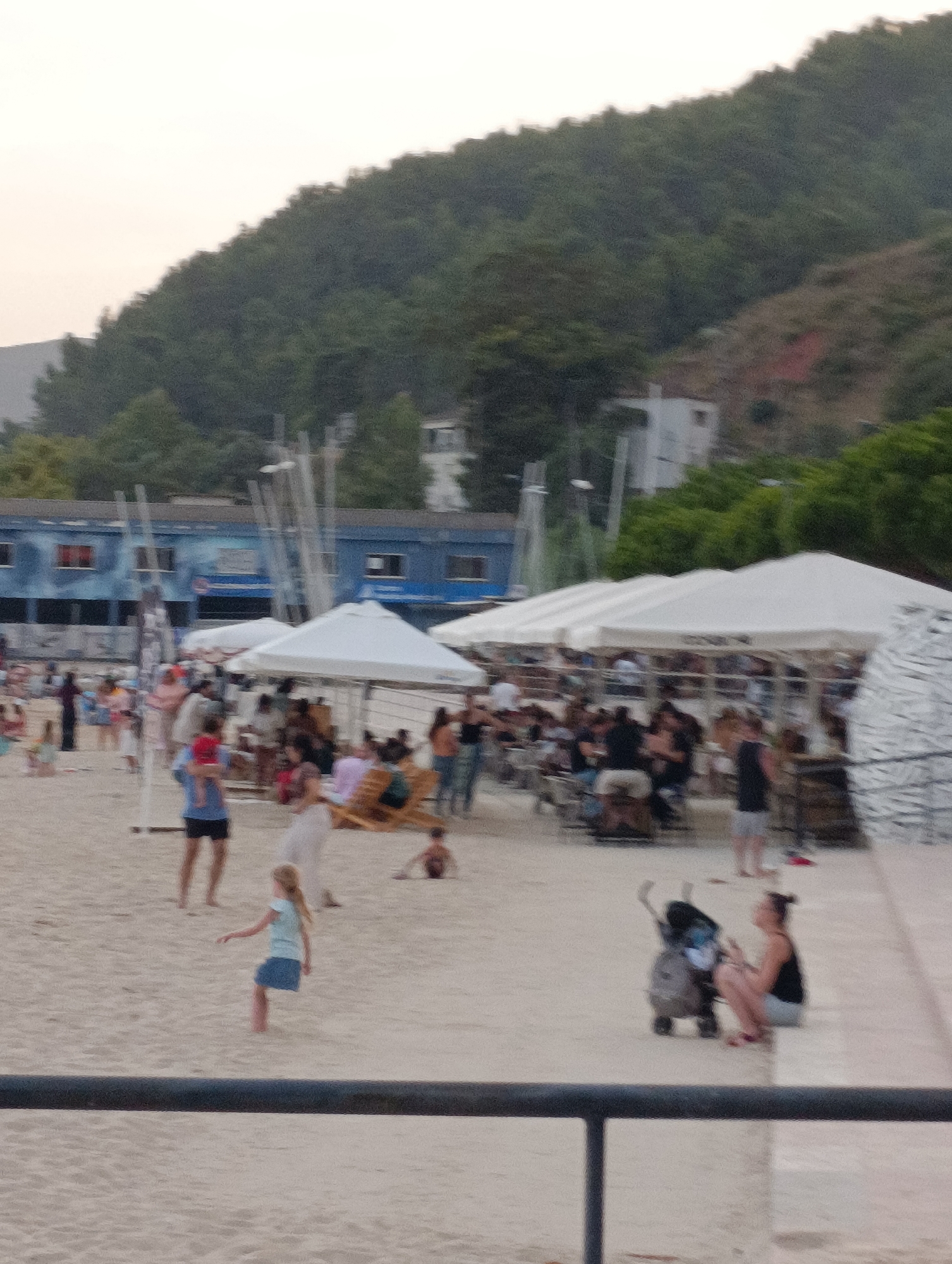 This media file captures a bustling beach scene, likely in Setúbal, Portugal. The setting is a sandy beach with a gentle slope leading to the water's edge, though the water itself is not visible. In the background, a lush green hillside forms a backdrop, dotted with some structures, including a blue building with windows and what appears to be a white building on the hill.
The foreground and midground are populated with numerous people enjoying a day at the beach. Several white tents are set up, providing shade for beachgoers, suggesting a commercial area or a designated gathering spot. People are seen sitting, standing, and walking on the sand.
Visible activities include socializing, with groups of people clustered together, and individuals relaxing under the tents. There's a child in a light blue top and dark skirt near the center of the image, appearing to be playing or dancing. Another young child, possibly being held by an adult, is visible towards the left. A woman is seated on a low wall near a stroller in the right portion of the image. The presence of a stroller and children suggests a family-friendly atmosphere.
The lighting indicates it might be late afternoon or early evening, with a soft, diffused light. The sky is overcast or hazy, and the overall atmosphere seems relaxed and casual. The people are dressed in casual beach attire. No discernible text is visible in the image. The scene conveys a sense of leisure and community on a warm day.