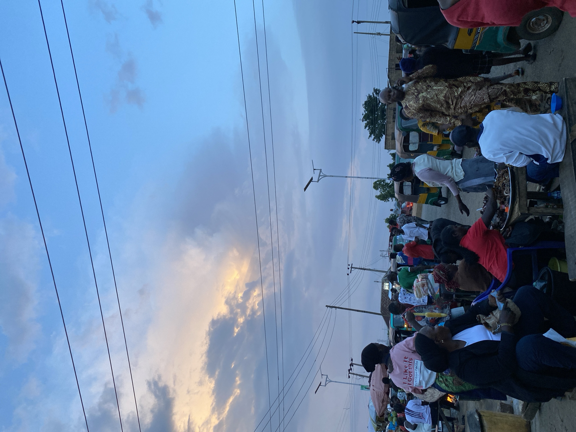 The image depicts a bustling outdoor market scene in Bukuru, Nigeria, likely during the late afternoon or early evening. The sky is a mix of blue with dramatic clouds, some illuminated by a warm sunset glow. Several power lines crisscross the sky above.
In the foreground and midground, a variety of people are engaged in market activities. A woman wearing a black jacket and white shirt is seated, holding a bag. Another woman in a red shirt is also seated, with food displayed on a table in front of her. A man in an ornate patterned garment is standing and speaking on a mobile phone. Other individuals are visible browsing goods, conversing, and moving through the market.
Various objects are present, including market stalls with awnings, stacked produce, what appear to be food items on tables, and several three-wheeled motorized vehicles commonly known as "Keke NAPEPs" or "Marwa" used for transportation and commerce. A solar-powered street light is visible on a pole. The ground appears to be unpaved. The overall impression is one of vibrant local commerce and daily life.