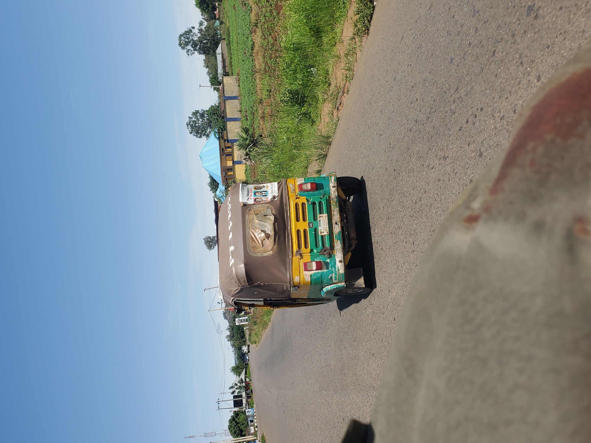 This media file shows a three-wheeled vehicle, commonly known as an auto-rickshaw or "keke," traveling on an asphalt road. The vehicle is predominantly yellow and green, with a brown fabric canopy. Visible on the rear of the auto-rickshaw are advertisements with images of people and text. The road appears to be in a rural or semi-urban setting, with a grassy field and some buildings on the left side of the road. Trees are also present in the background. The sky is clear and blue, suggesting it is daytime and the weather is fair. The camera perspective seems to be from another vehicle moving alongside or slightly behind the auto-rickshaw. The image is oriented vertically, with the sky at the left and the road and auto-rickshaw filling the right side. There are no discernible emotions conveyed, and the scene appears to be a typical day in a Nigerian location, likely Panyam, given the context.