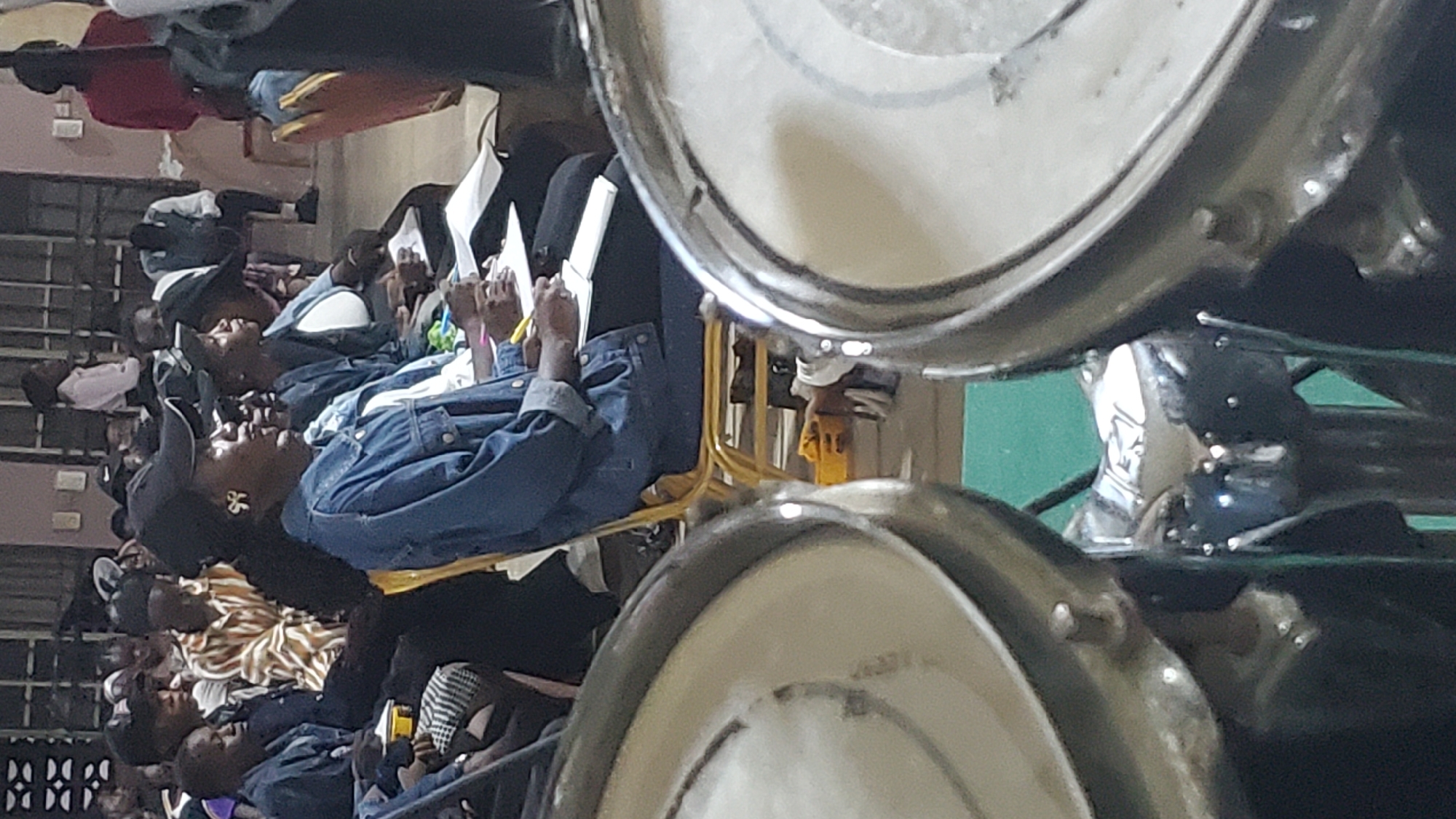 This media file depicts a group of people, likely attending an indoor event or gathering. The subjects are predominantly seen from the chest up, with many individuals appearing to be seated. They are mostly young adults, and their clothing suggests a casual or semi-formal setting. Some are looking down, possibly at papers or something in their laps, while others are looking forward or slightly to the side, engaged with their surroundings.
Notable details include several individuals wearing denim jackets, suggesting a possibly cool indoor temperature or a fashion choice. One person in the foreground is wearing a black baseball cap and large hoop earrings. Another individual is wearing a striped shirt. The presence of what appear to be large, metallic objects, possibly drums or part of a musical instrument, in the foreground and partially obscuring the view, indicates that the event might involve music or performance. The lighting appears to be artificial, consistent with an indoor setting.
The scene is likely taking place in Jalingo, Nigeria, as indicated by the location context. There are no clear indicators of the specific time of day or weather conditions. The overall mood seems attentive and engaged, with no overt signs of distress or excitement. The arrangement of people suggests a seating area, perhaps for an audience or participants in a workshop or meeting. No visible text is present in the image.