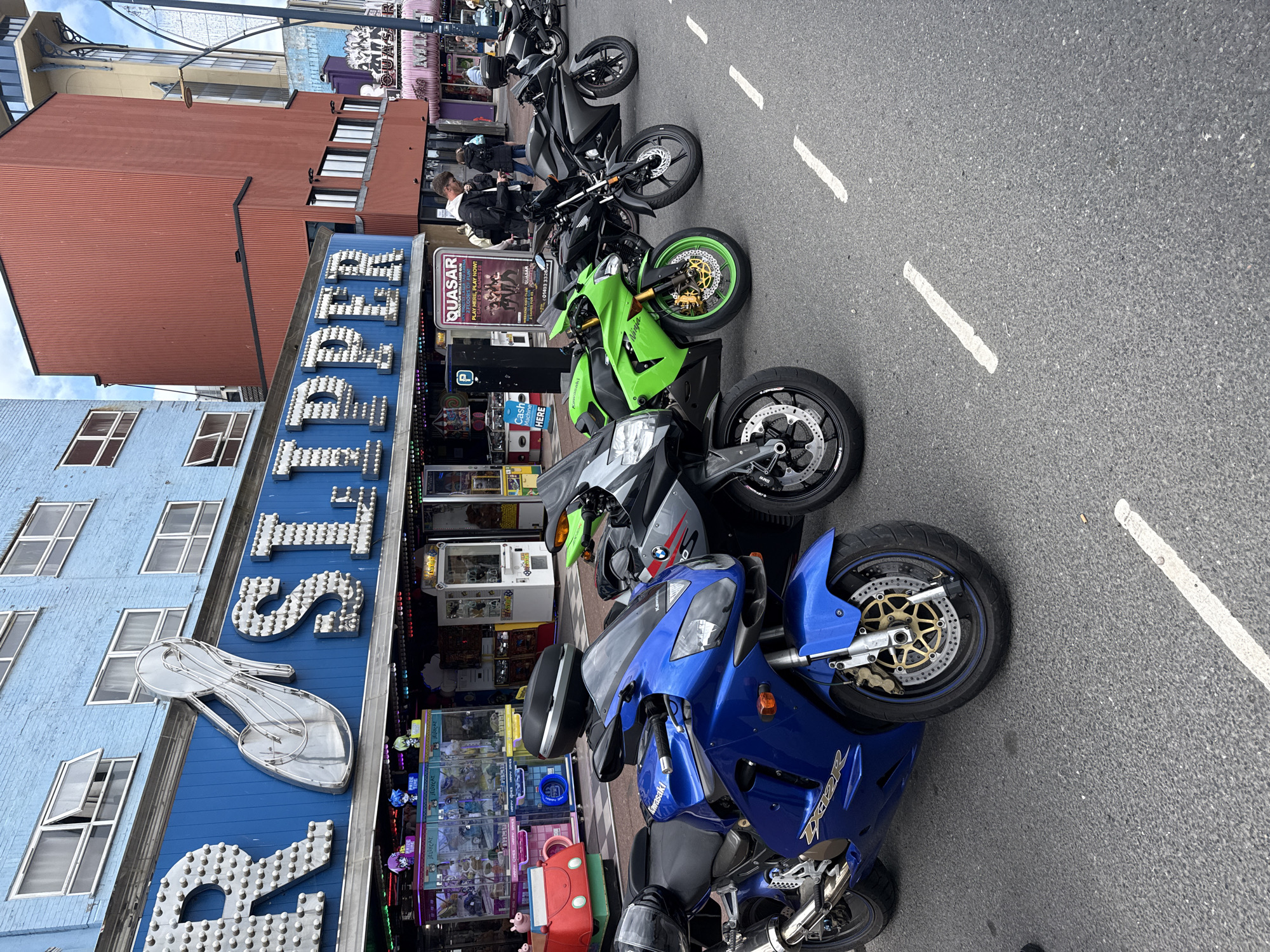 Here's a detailed description of the image:
**Scene:** A row of motorcycles parked along a street next to a building in what appears to be a commercial district.
**Location:** Based on the presence of "Jellycat" and "Quasar" signs, this appears to be a street in Great Yarmouth, United Kingdom. These businesses are located on the main strip in the city.
**Objects & Environment:**
* A blue building with the word "Jellycat" in large, lit-up letters and a neon shoe sign. The building features several windows.
* A Quasar sign is visible in front of a reddish-brown building, advertising gaming.
* There are several arcade-style game machines visible.
* The street is paved and has painted white lines marking lanes.
**Subjects:**
* Multiple motorcycles are parked in a line. They are various colors, including blue, silver, green, and black.
* There are people visible, possibly looking at the bikes or entering the building.