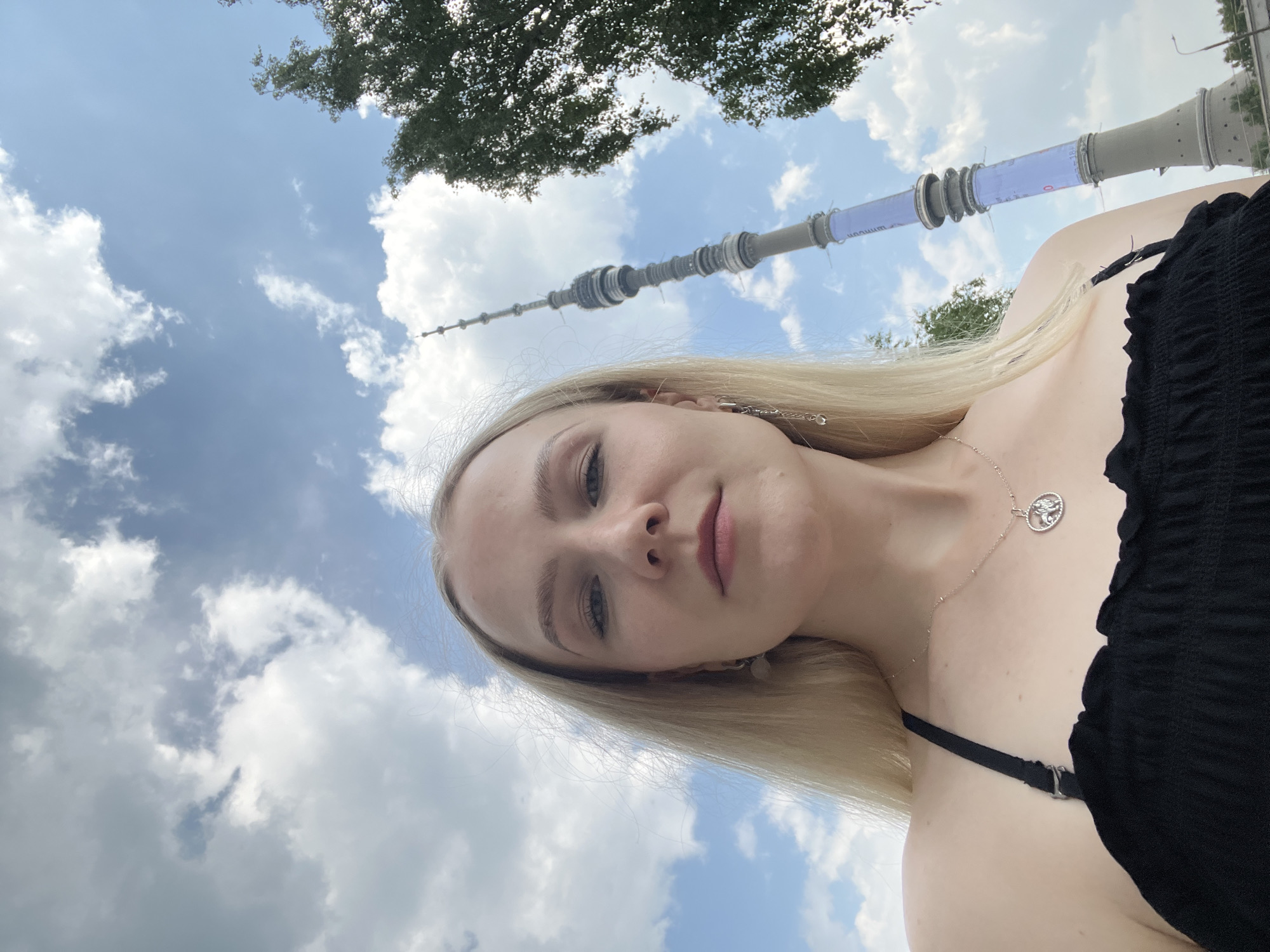A young woman with blonde hair is taking a selfie outdoors. She is wearing a black top and a silver necklace with a round pendant. In the background, there is a tall tower, a tree, and a cloudy sky. The tower appears to be the Ostankino Tower, which is located in Moscow, Russia, not Khimki. The image captures a casual, daytime scene with a focus on the woman and the prominent landmark in the background.