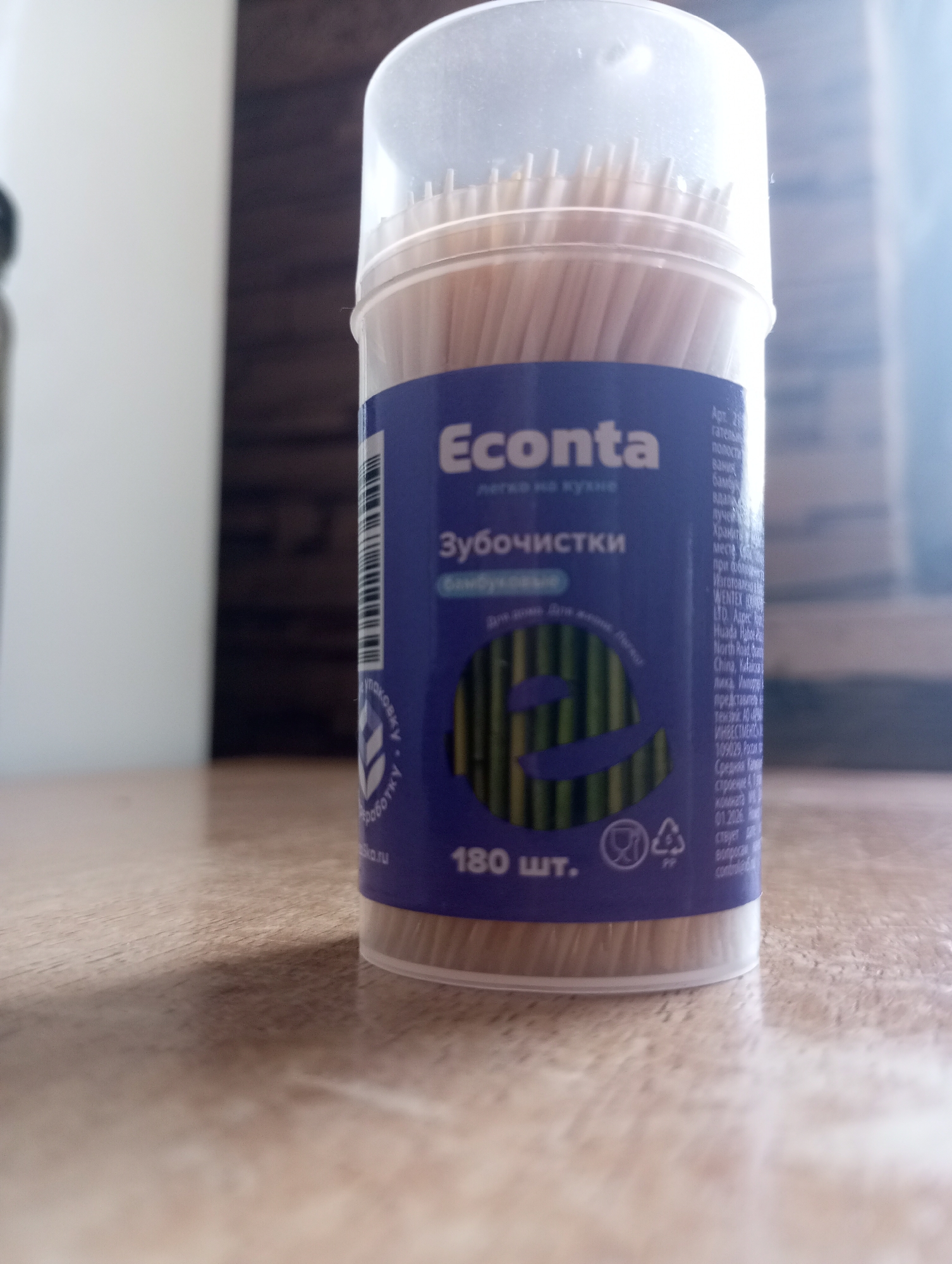 A close-up, low-angle, eye-level shot shows a transparent plastic container filled with bamboo toothpicks. The container has a blue label with text and a logo that appears to be a stylized letter "E" formed by bamboo stalks. The word "Econta" is prominently displayed on the label in white text, along with "Зубочистки" (toothpicks). Below this, it says "для дома. для кухни." (for home. for kitchen.). The bottom of the label indicates "180 шт." (180 pcs.) and has recycling symbols. The container is sitting on a light brown, textured surface that appears to be a table or countertop. The background is out of focus, showing a dark, wooden-textured surface and a bright, possibly window-lit area on the right. There are no people or discernible actions in the image. The overall scene is indoors, likely a domestic or retail setting, with no specific indicators tying it to Kirzhach, Russia.