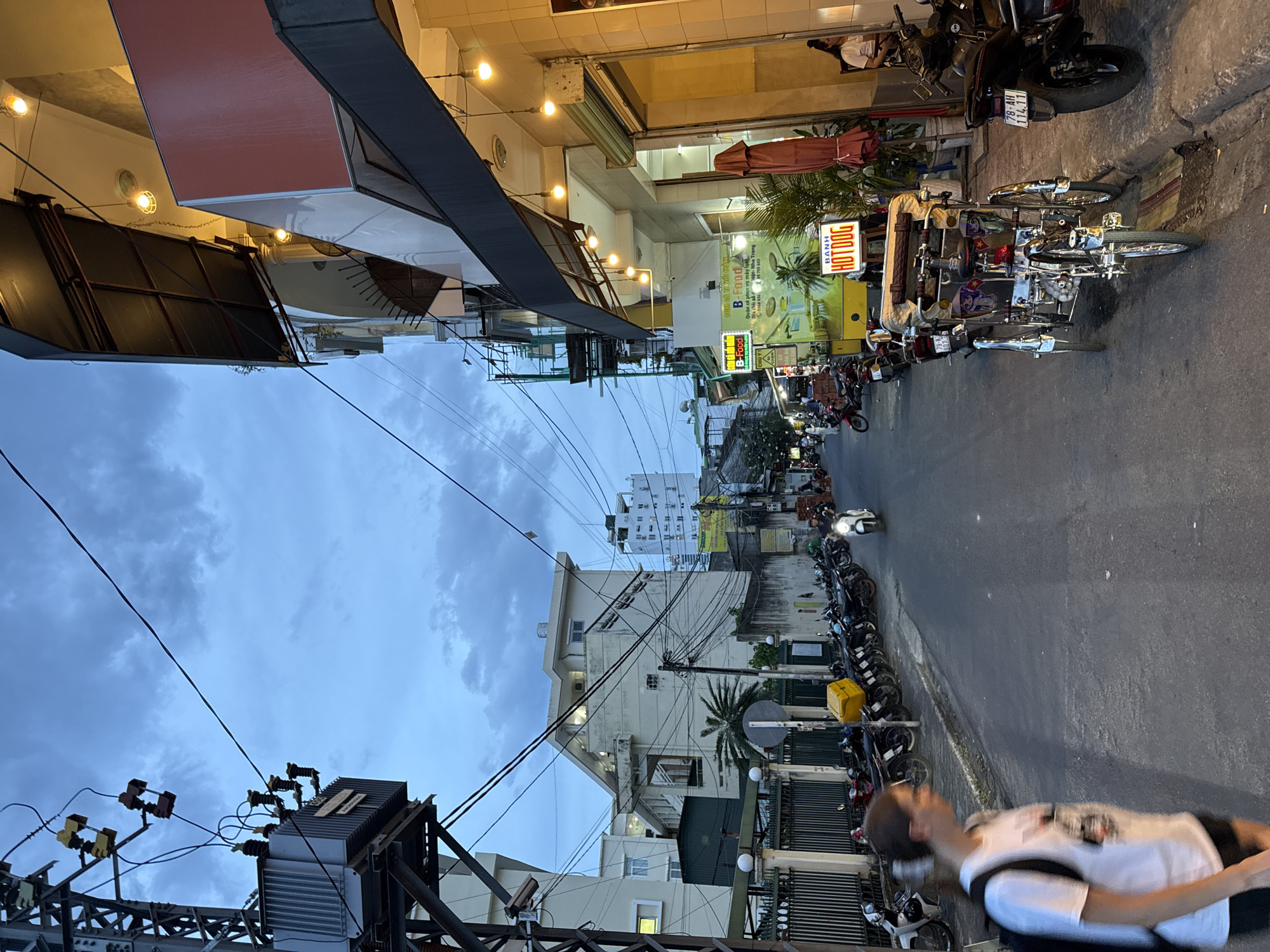 The image depicts a street scene in Nha Trang, Vietnam, at dusk. The sky is overcast with dark clouds. On the left side of the street, a large electrical transformer is mounted on a metal pole, with numerous wires extending from it. A person wearing a white t-shirt and dark shorts, with a backpack, is walking away from the camera, partially blurred. Adjacent to the transformer pole is a metal fence. A row of parked motorcycles lines the right side of the street.
On the right side of the street, a building features a covered outdoor seating area with hanging lights. A sign for "B FOOD" and "BANH HOTDOG" is visible, indicating food establishments. Another building further down the street appears to be a multi-story residential or commercial structure. Several more motorcycles are parked along the sidewalk. A three-wheeled vehicle, possibly a cyclopousse or pedi-cab, is parked on the side of the road. In the background, taller buildings are visible, and a person on a white scooter is riding down the street. The street is paved with asphalt.