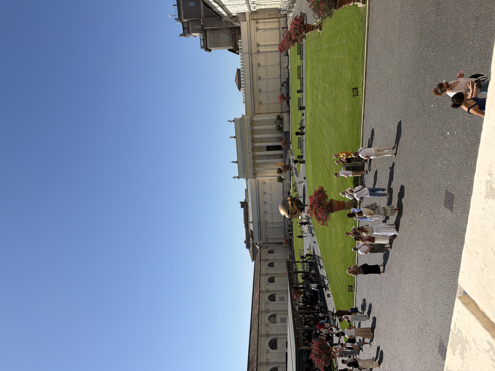 This is a wide-angle, outdoor, eye-level shot of the Pinacoteca Vaticana courtyard in Vatican City. The sky is a clear, bright blue. In the center of the frame is a large, golden, spherical sculpture, "Sphere within Sphere" by Arnaldo Pomodoro. The sculpture is positioned on a green, manicured lawn surrounded by benches and potted plants with red flowers.
To the left of the lawn is a covered outdoor seating area with tables and chairs, populated by many people. A sign for "Bistrò" is visible on one of the structures. Behind this area, and extending to the right, are long, pale yellow buildings with arched windows and classical architectural elements. The building in the center features a prominent portico with columns and statues.
The foreground is paved with a cobblestone-like pattern and is populated by a group of tourists. Most are standing, some are looking at the sculpture, others are talking amongst themselves, and a few are walking. They are dressed in casual summer attire. Two women are walking towards the viewer in the lower right corner of the frame, one in a blue top and the other in a white top and pink skirt.