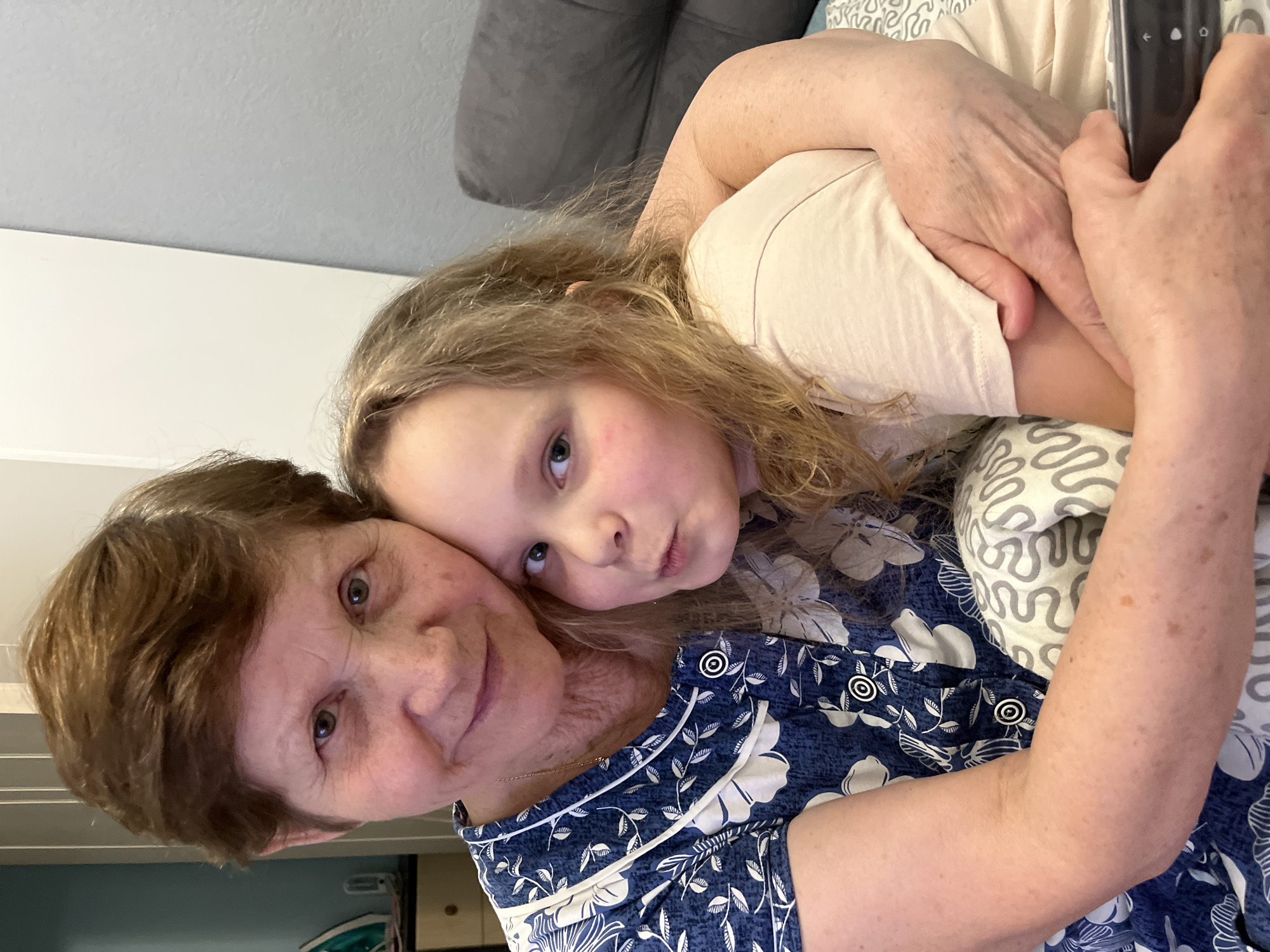 An elderly woman with short brown hair and a young fair-haired child are tightly embracing in an indoor setting. The woman is wearing a dark blue floral patterned shirt. The child is wearing a light beige t-shirt. A remote control is visible in the upper right corner of the image. Behind them is a grey sofa and patterned pillows. The location or whether it is in the city of Izhevsk, Russia, cannot be determined from the image.