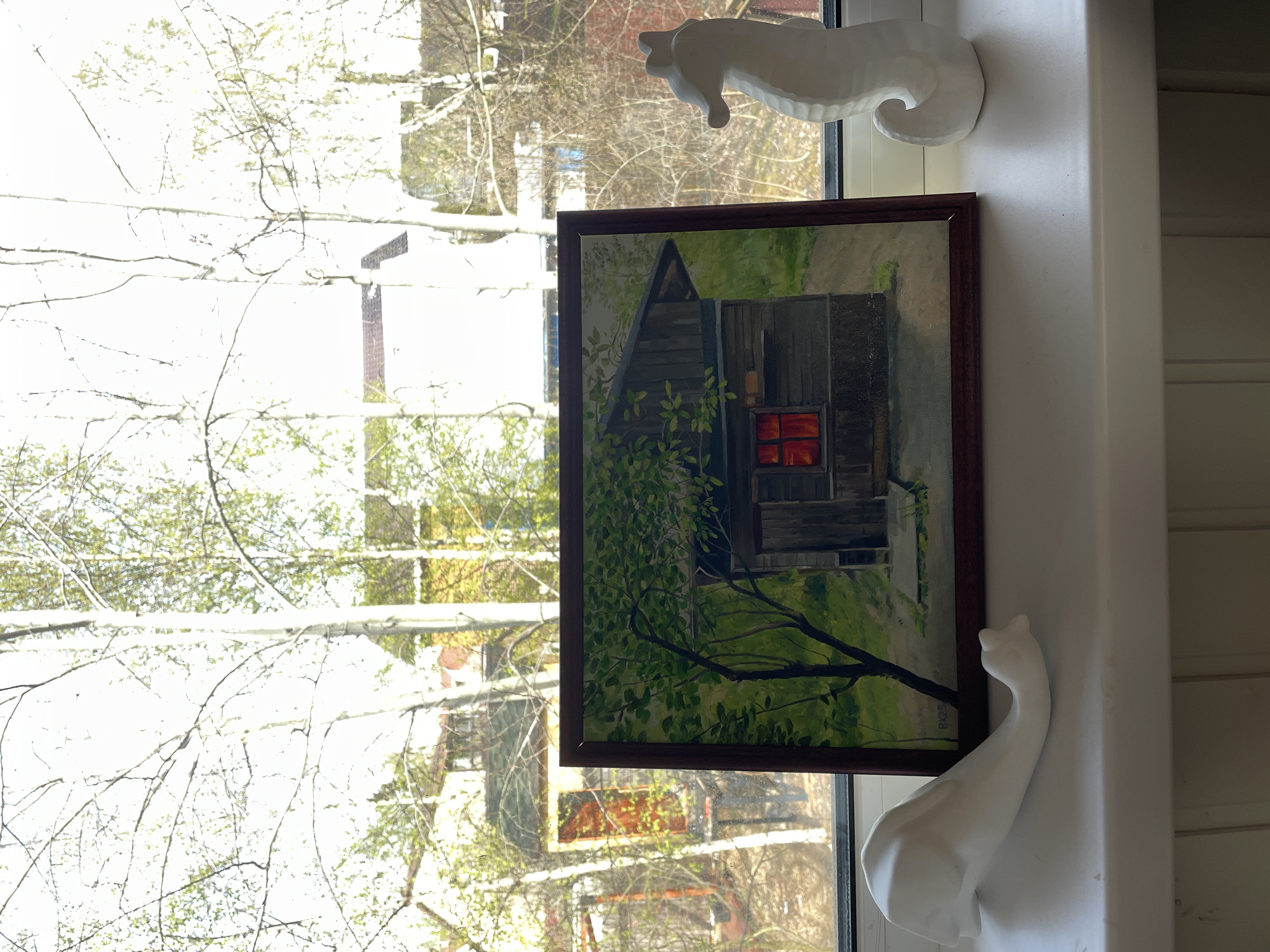 The image shows a framed painting of a cabin hanging on a wall, positioned near a window. Outside the window, trees with budding leaves are visible. A white ceramic seahorse figurine sits on the windowsill above the painting, while a white ceramic swan figurine sits below it. The scene appears to be indoors, possibly a home in Sestroretsk, Russia, although there is no definitive information in the image that confirms this location. The interaction is between the artwork and the decorative items enhancing the domestic setting.