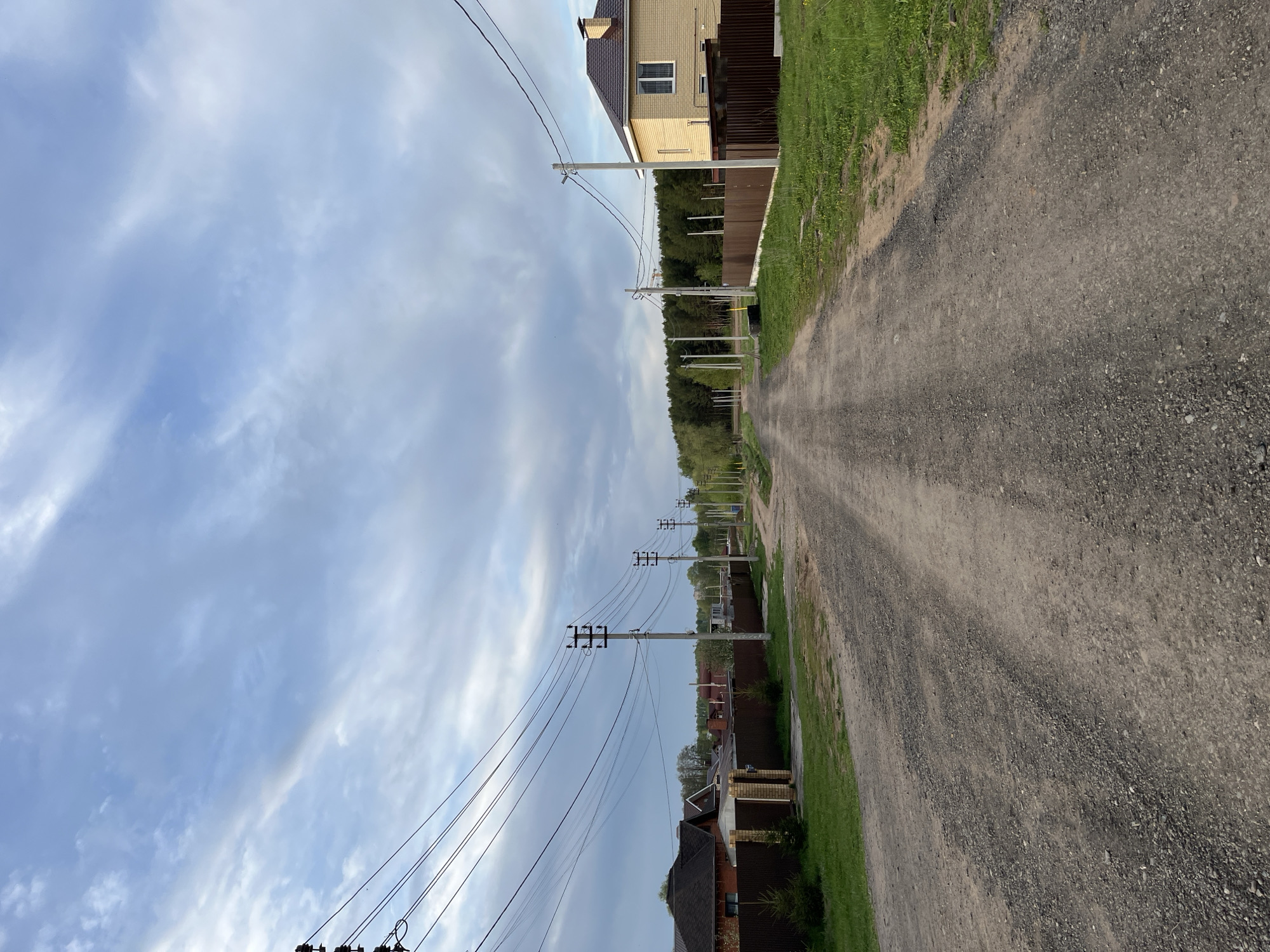 A ground-level, medium shot shows a straight gravel road extending into the distance. The photo, taken in Chaikovsky, Russia, is vertically oriented. The road is flanked by fences, houses, and trees. Power lines and poles run along the left side of the road, contrasting against a cloudy sky. The perspective suggests a rural or suburban setting.