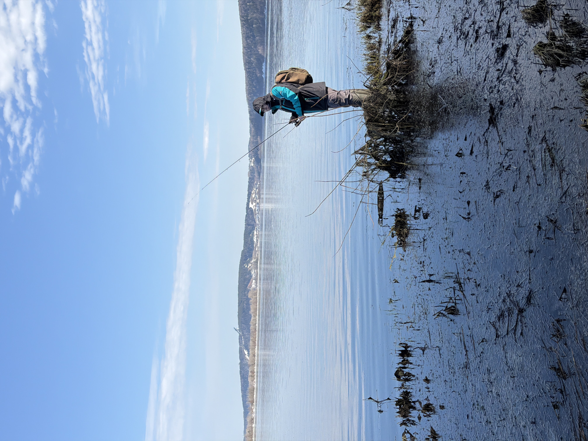 An outdoor scene in Unknown, Unknown, shows an individual fishing in a shallow, partially frozen or slush-covered body of water under a clear blue sky. The individual, an adult of indeterminate gender, is positioned right of center, wearing a black and teal hooded jacket with the hood up, dark pants, dark knee-high boots, and dark gloves. A light brown backpack with multiple compartments is worn on their back. They are holding a fishing rod with a reel, extending the rod tip over the dark water. The immediate foreground water is dark and shallow, displaying intricate patterns of thin ice or slush formations on its surface, interspersed with dense clusters of dark, submerged or emergent plant debris and branches where the person stands. The main body of water beyond is calm, reflecting the blue sky, and extends to a distant horizon. On this horizon, a long, low-lying forested hill range is visible, featuring distinct patches of snow and a prominent dark structure, possibly a ski lift or tower, on one of the snowy slopes. The sky above is clear, light blue, with scattered, wispy white clouds.