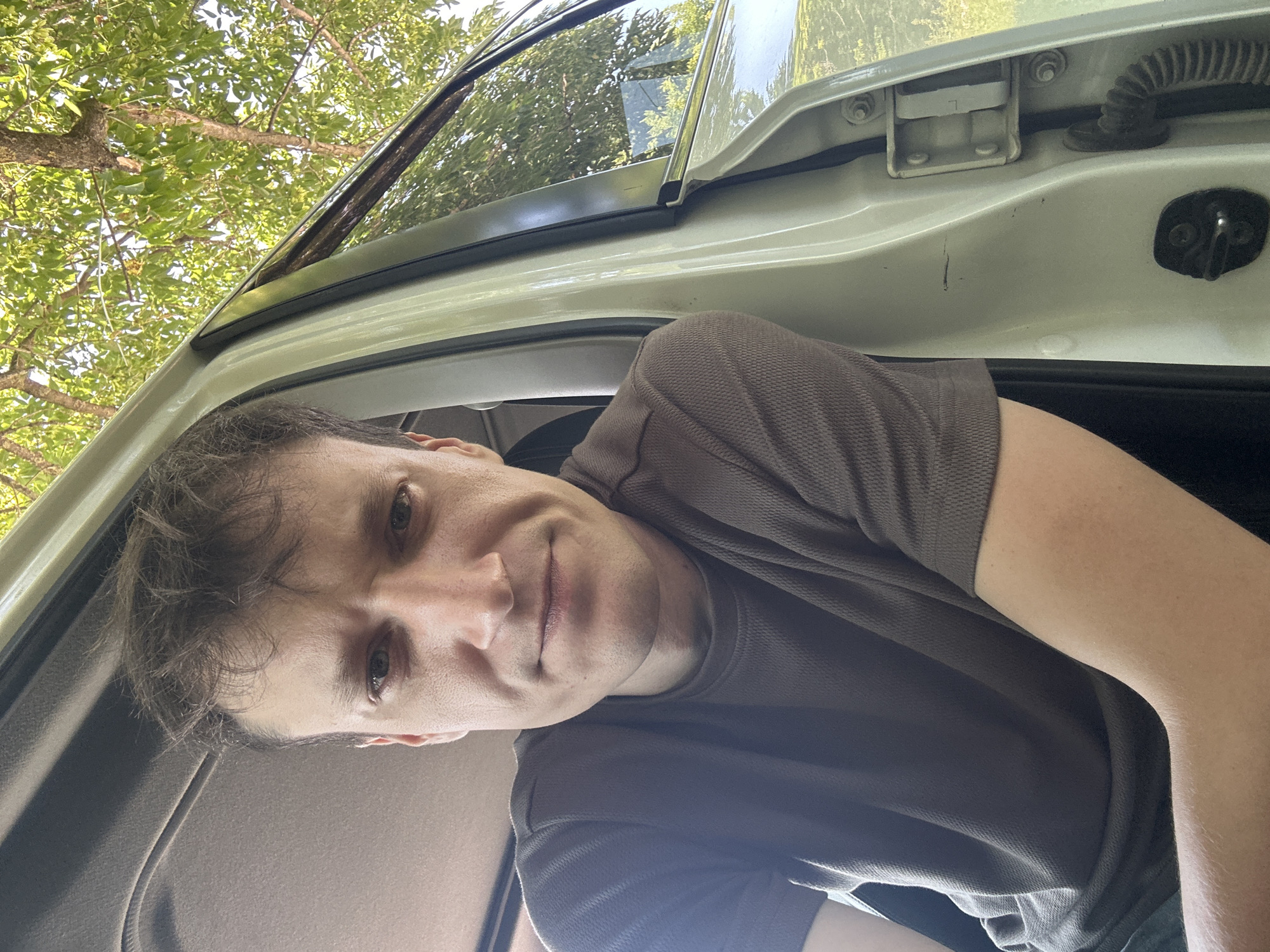 A close-up, chest-up selfie shows a person with light skin, brown hair, and a dark brown t-shirt. The person is inside a car, with the passenger door open. The car's interior is visible, as is the exterior environment. Sunlight filters through the trees outside, creating dappled light. The car appears to be parked on a dirt road. The image is likely taken in the city of Makiv, Ukraine, given the context provided.
