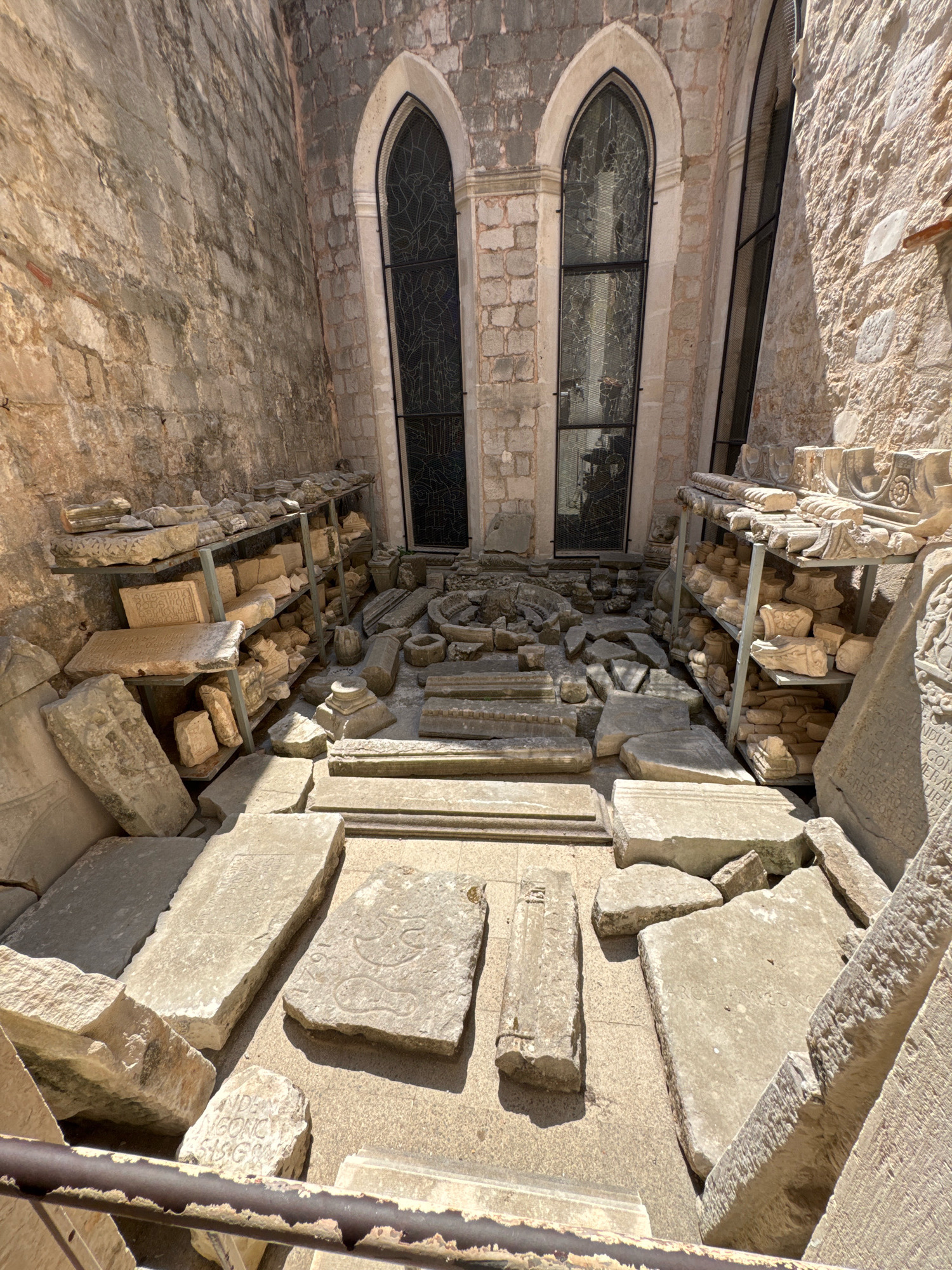 The image shows an outdoor courtyard with ancient stone fragments displayed on shelves and on the ground. The courtyard is enclosed by stone walls, and two tall, arched windows with dark glass are visible in the background. The stone fragments appear to be architectural elements and gravestones, some with inscriptions and carvings. The scene is lit by natural sunlight, casting shadows on the ground. This courtyard is located in the Rector's Palace in Dubrovnik, Croatia.