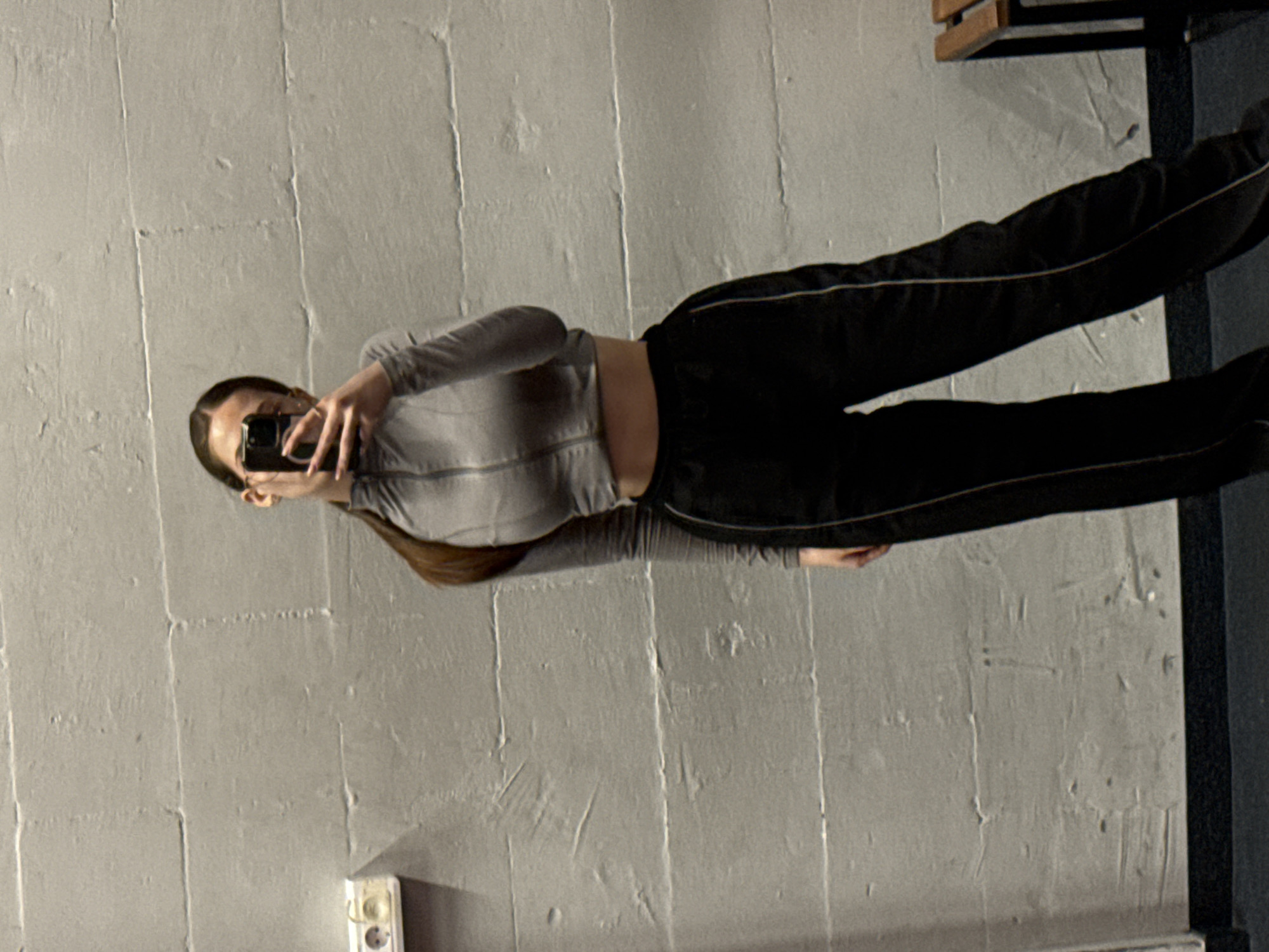 A woman is taking a selfie in a room with light gray walls. She is wearing a long-sleeved gray crop top and black pants with a white stripe down the side. Her hair is brown and pulled back. A partially visible electrical outlet is on the wall near the bottom of the frame. A piece of furniture is visible in the upper right corner. The location is unknown and there is no clear connection to Sofia, Bulgaria.