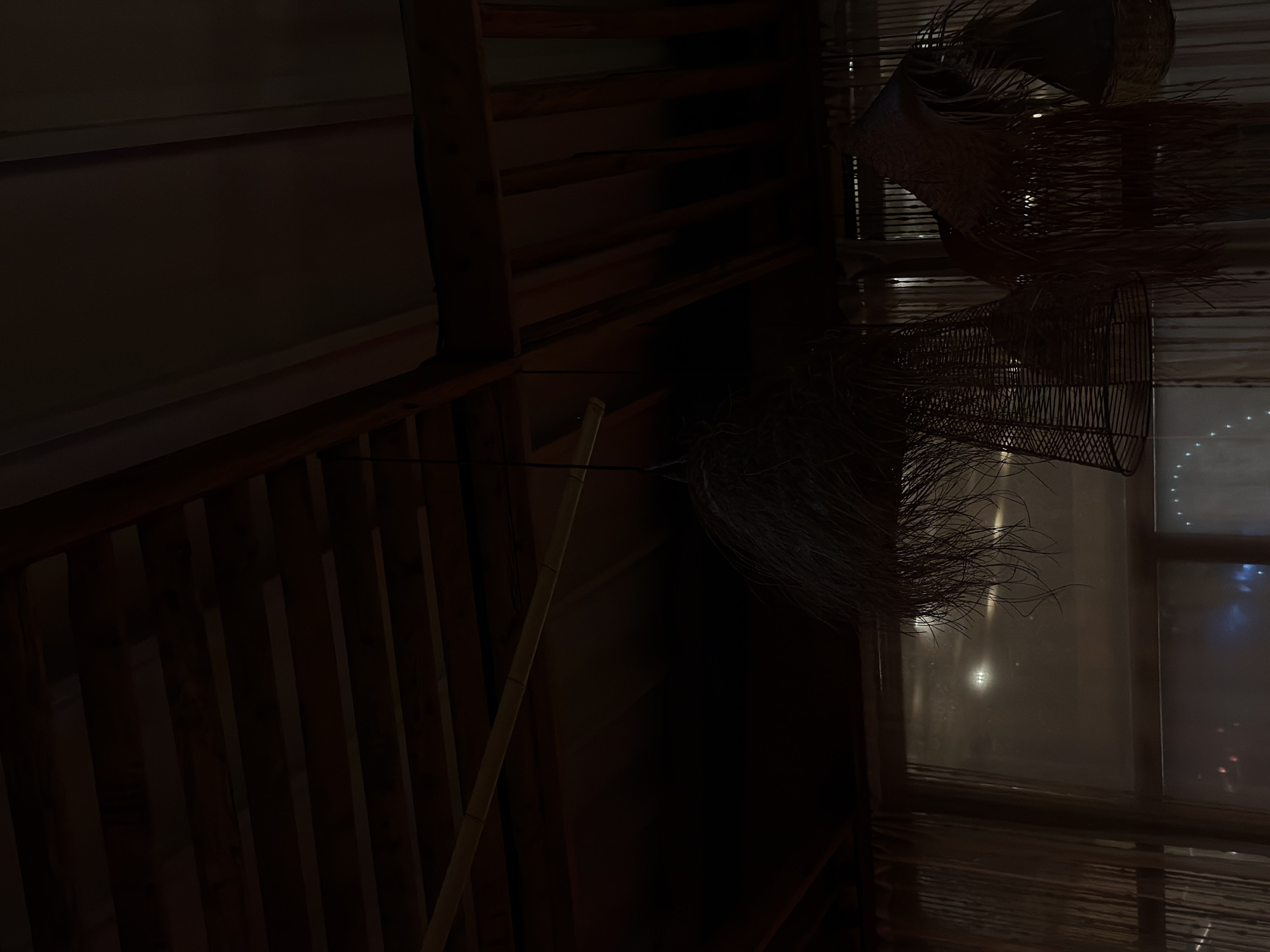 This appears to be an interior shot of a building, possibly in Yerevan, Armenia, although there's no direct evidence to confirm this location. The image is dark, with a heavy emphasis on wood, visible in beams and slats suggesting either a ceiling or a wall structure. There is a hint of outdoor light coming through what appears to be a window or opening covered with a semi-transparent material. Woven or wicker-like decorative items hang in front of the window.