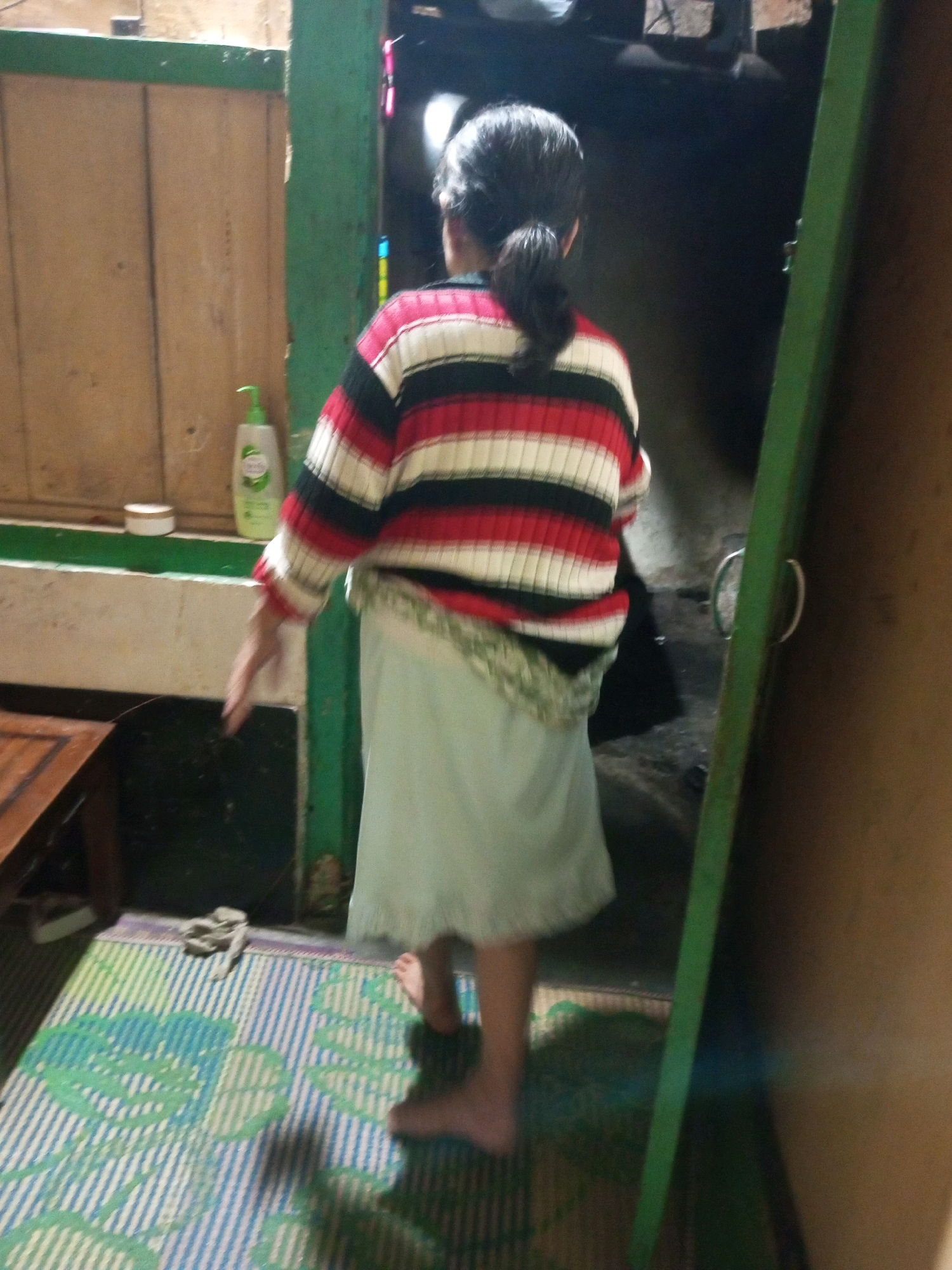 A person, viewed from behind, is standing in a doorway. They are wearing a striped sweater with horizontal bands of red, white, and black, and a light blue skirt. The person's hair is pulled back into a ponytail. To the left of the person, on a shelf, are two items: a white circular container and a green plastic bottle with a pump dispenser. The background is dimly lit and appears to be an interior space. The floor is covered by a blue and white striped mat with green patterns. A wooden table is partially visible on the left side of the frame. The overall scene suggests a domestic setting in Andongsili, Indonesia.