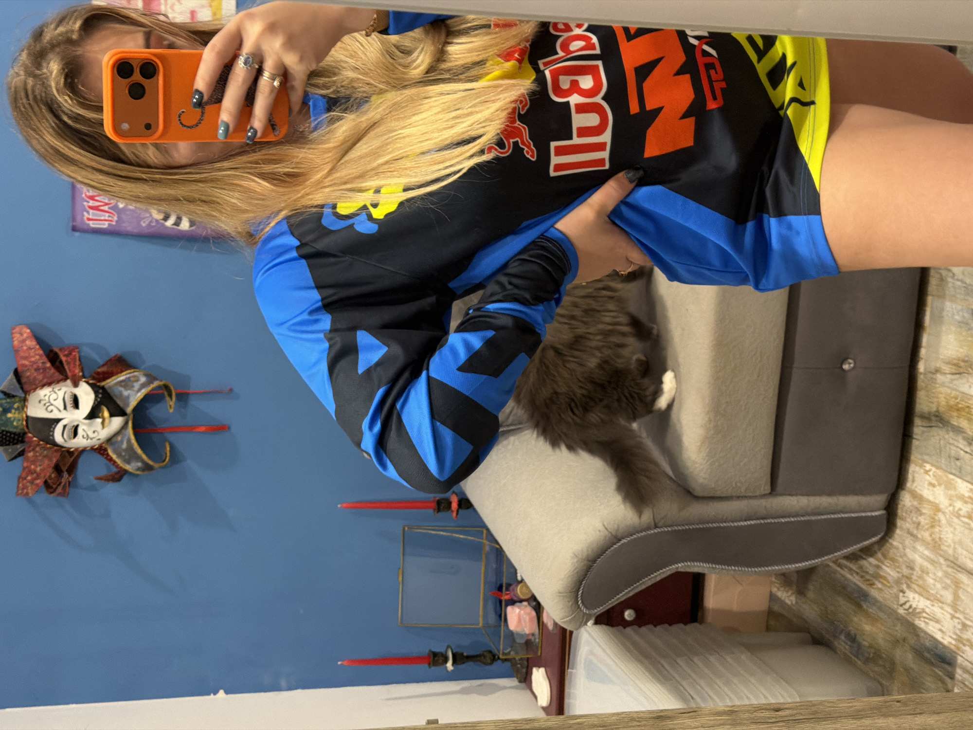 A woman is taking a selfie in what appears to be a reflection in Большая Шильна, Russia. She is holding an orange iPhone with a tiger case. The woman has long, blonde hair and is wearing a racing-style shirt with "Red Bull" branding, along with blue and yellow accents. Her nails are painted dark with glitter.
Behind her, a blue wall is visible, decorated with a mask and candles. The woman is next to a grey sofa on which a grey cat sits. A cabinet with decorative objects is visible below the wall decorations.