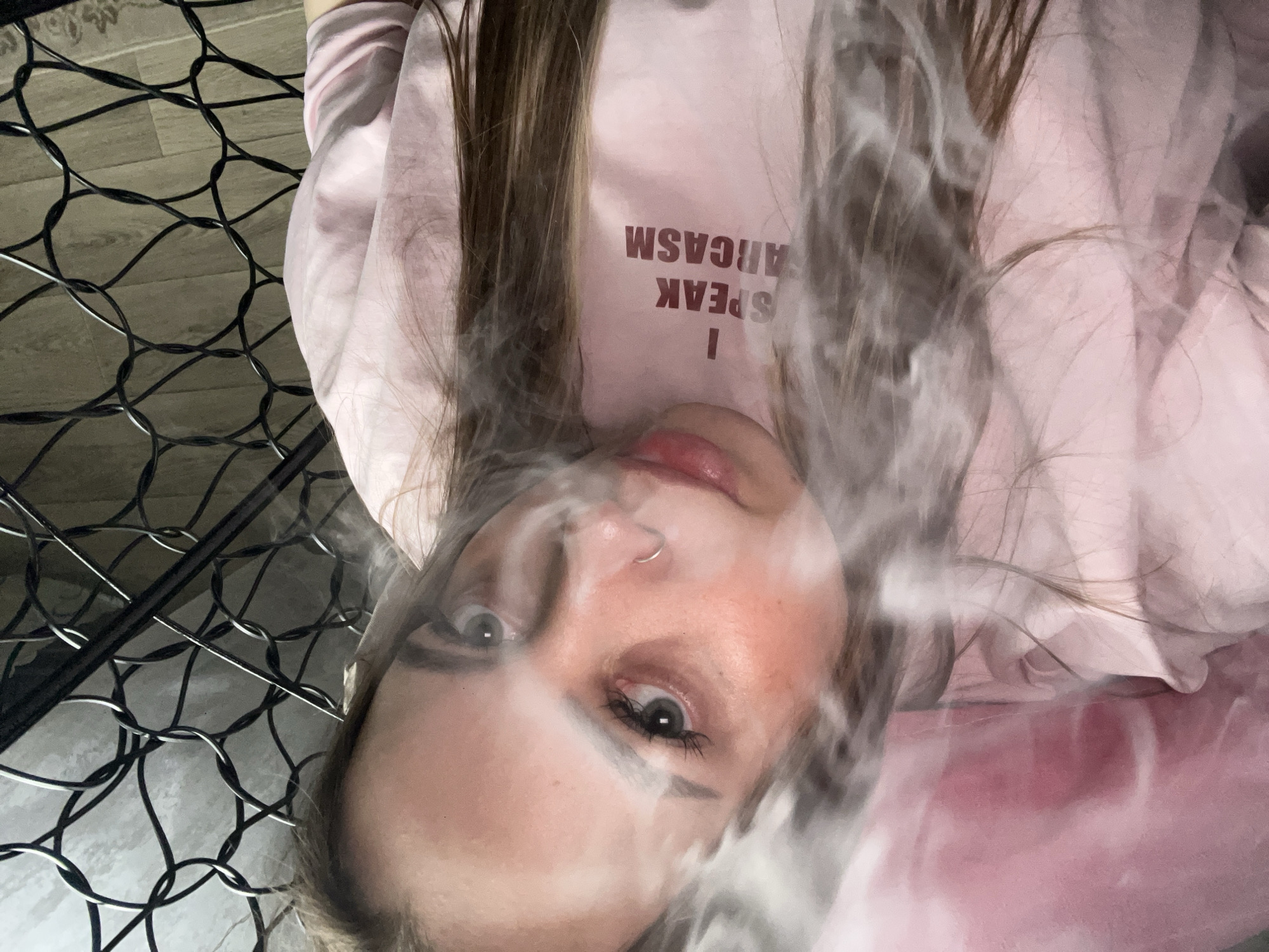 A woman is pictured indoors with gray smoke obscuring her face. She has long brown hair and a nose ring. She wears a light pink sweatshirt with the words "I SPEAK SARCASM" printed on it upside down. The background features a floor made of light-colored wood and a black wire mesh structure. The location is not mentioned in the image.