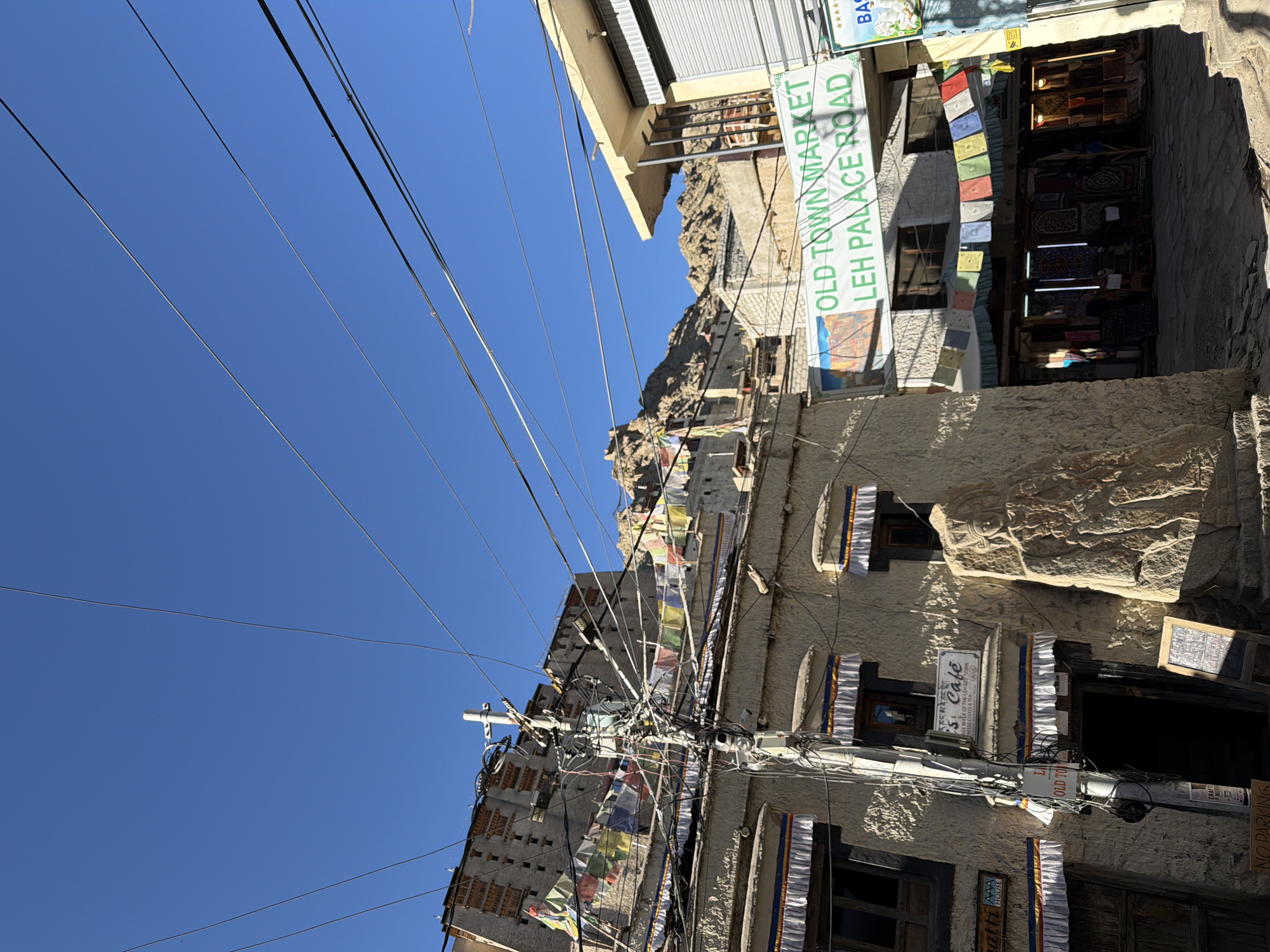 The image captures a view of a street in Leh, India, with a clear blue sky overhead. In the foreground, a building with traditional architecture is visible, featuring wooden window frames and decorative fabric borders above the windows. A utility pole heavily laden with wires stands in front of this building. Prayer flags are strung across the scene, adding a splash of color against the muted tones of the buildings and the landscape.
Further back, a multi-story structure resembling a palace or fort is perched on a rocky hillside, a common sight in Leh. To the right, another building with a modern awning is present, displaying a sign that reads "OLD TOWN MARKET LEH PALACE ROAD". Below this sign, a marketplace appears to be set up with stalls displaying various textiles. The overall scene depicts a blend of traditional and contemporary elements characteristic of Leh, a city known for its Buddhist heritage and mountainous surroundings. There are no visible people in the image.