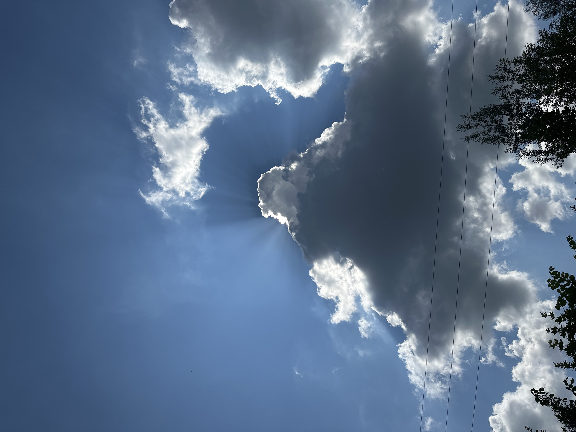 The image depicts a cloudy sky with crepuscular rays emanating from behind a large, dark cloud. The sky is a vibrant blue, with smaller, white clouds scattered throughout. In the foreground, there are silhouetted tree branches and power lines. The overall scene is a natural, outdoor environment. The location is specified as the city of Nizhny Novgorod, Russia. There are no people or animals visible in the image.