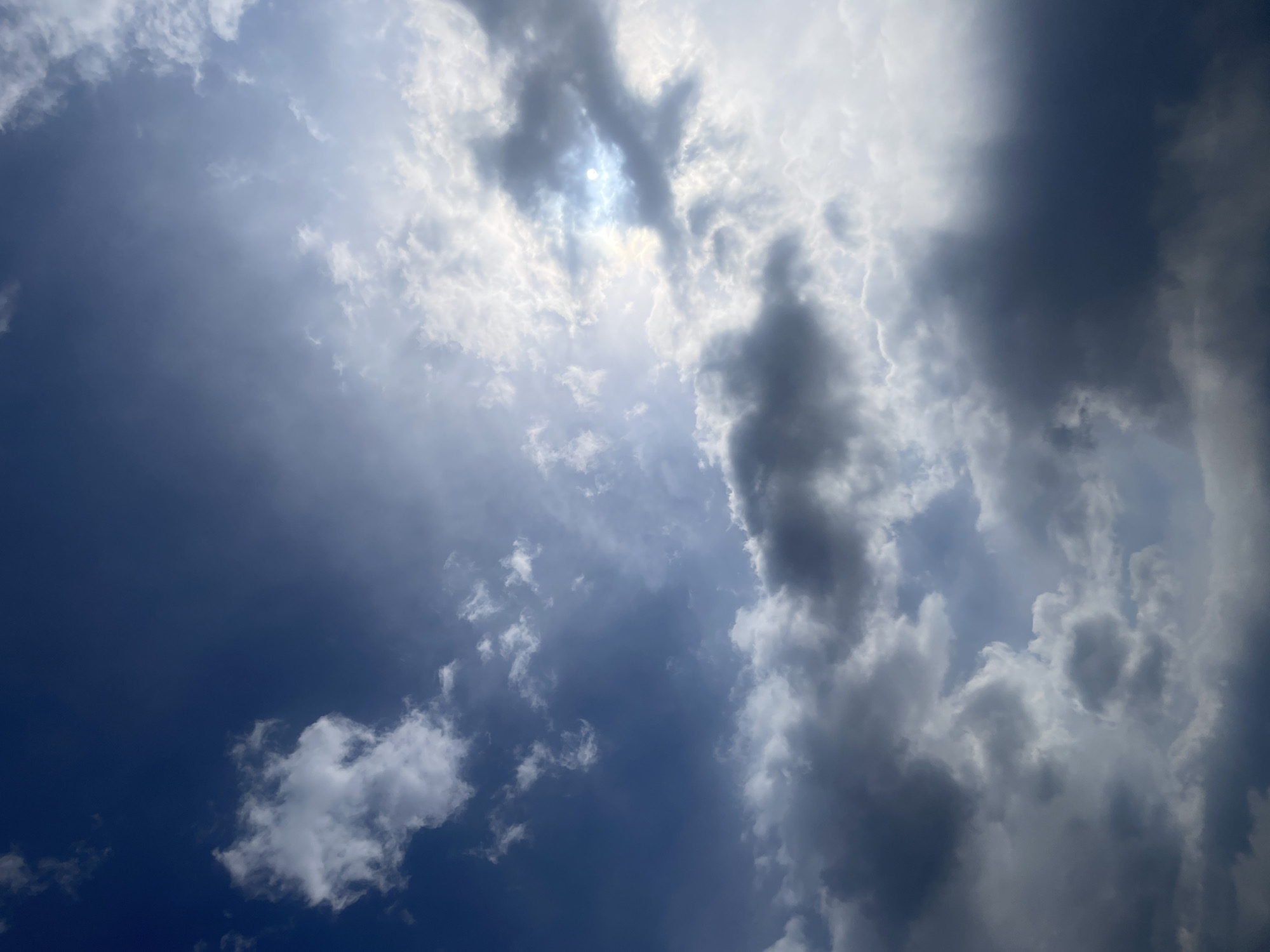 The media file presents an upward view of a dynamic sky filled with varying cloud formations and the sun. The primary subjects are the sky itself and the clouds.
The sky exhibits a gradient of colors, ranging from a deep, vibrant blue in the lower-left quadrant to lighter shades of blue, grey, and white where clouds dominate and the sun breaks through. The sun is visible in the upper-central portion of the image, partially obscured by bright, luminous white clouds, creating a strong, radiant glow. This bright area suggests intense sunlight piercing through the cloud cover.
Clouds are abundant and diverse. Towards the right side and upper-right, darker, denser grey clouds are prominent, indicative of thicker atmospheric conditions. In contrast, the lower-left and central areas feature more scattered, wispy white clouds set against the bluer portions of the sky. Rays of light can be observed emanating from the sun's position, illuminating the textures of the surrounding clouds.
No people, man-made objects, or any discernible text are visible within the frame, focusing solely on the natural atmospheric phenomenon. The scene captures a moment of fluctuating weather, likely a partly cloudy or clearing day. The intensity and high position of the sun suggest it is daytime, possibly around midday or early afternoon. The location context provided is Yerevan, Armenia, though no specific geographical landmarks are present in the image itself.