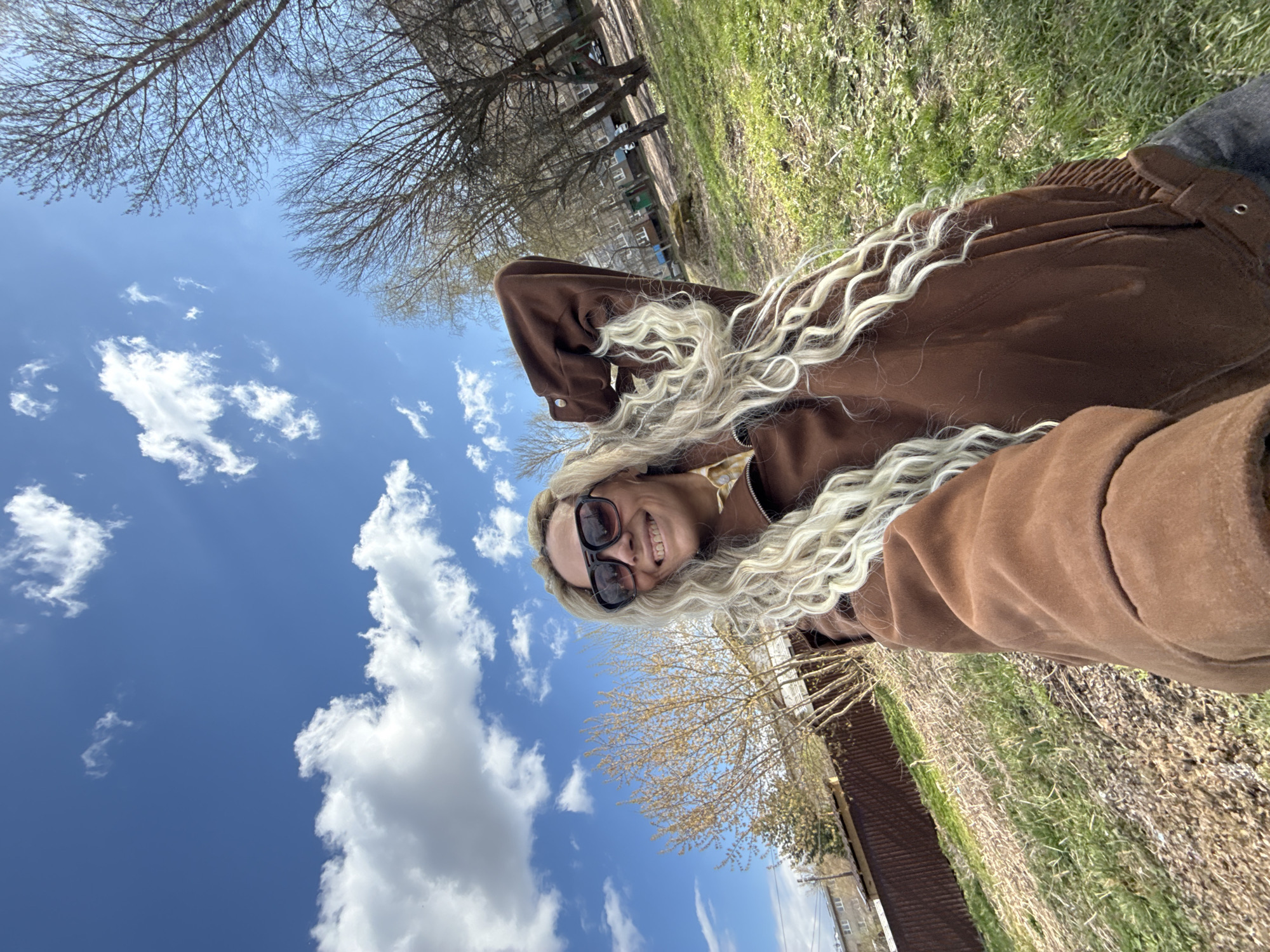 The image, rotated 90 degrees clockwise, features a person lying on green grass, taking a selfie. The subject is a woman with long, wavy blonde hair, wearing dark sunglasses and a brown jacket. She is smiling broadly, looking up towards the camera.
The background consists of a bright blue sky with numerous white, fluffy clouds. Silhouetted against the sky are the bare branches of several trees, indicative of late winter or early spring. In the distant background, multi-story residential buildings are faintly visible, suggesting an urban or suburban park setting.
The scene takes place during daylight hours, under clear and sunny weather. The woman's smile conveys a feeling of happiness and relaxation. No visible text is present in the image. The location is Rusanovo, Russia.