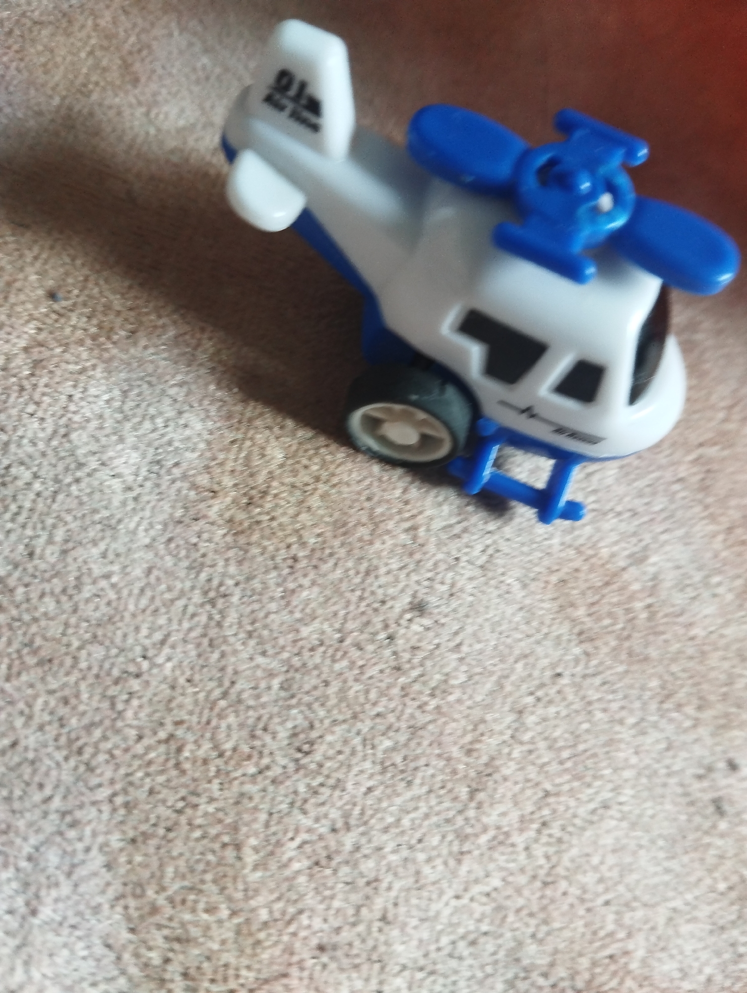 A toy helicopter, primarily white with blue accents, rests on a textured, light brown carpeted surface. The helicopter features a visible rotor on top, black windows, and two wheels. Text is present on the tail of the helicopter, appearing to read "911 Air Rescue". The image is taken from a slightly elevated angle, with the carpet filling most of the frame. No people or further environmental context are discernible. The location in Orlovka, Kyrgyzstan, is not visually represented in the image.
