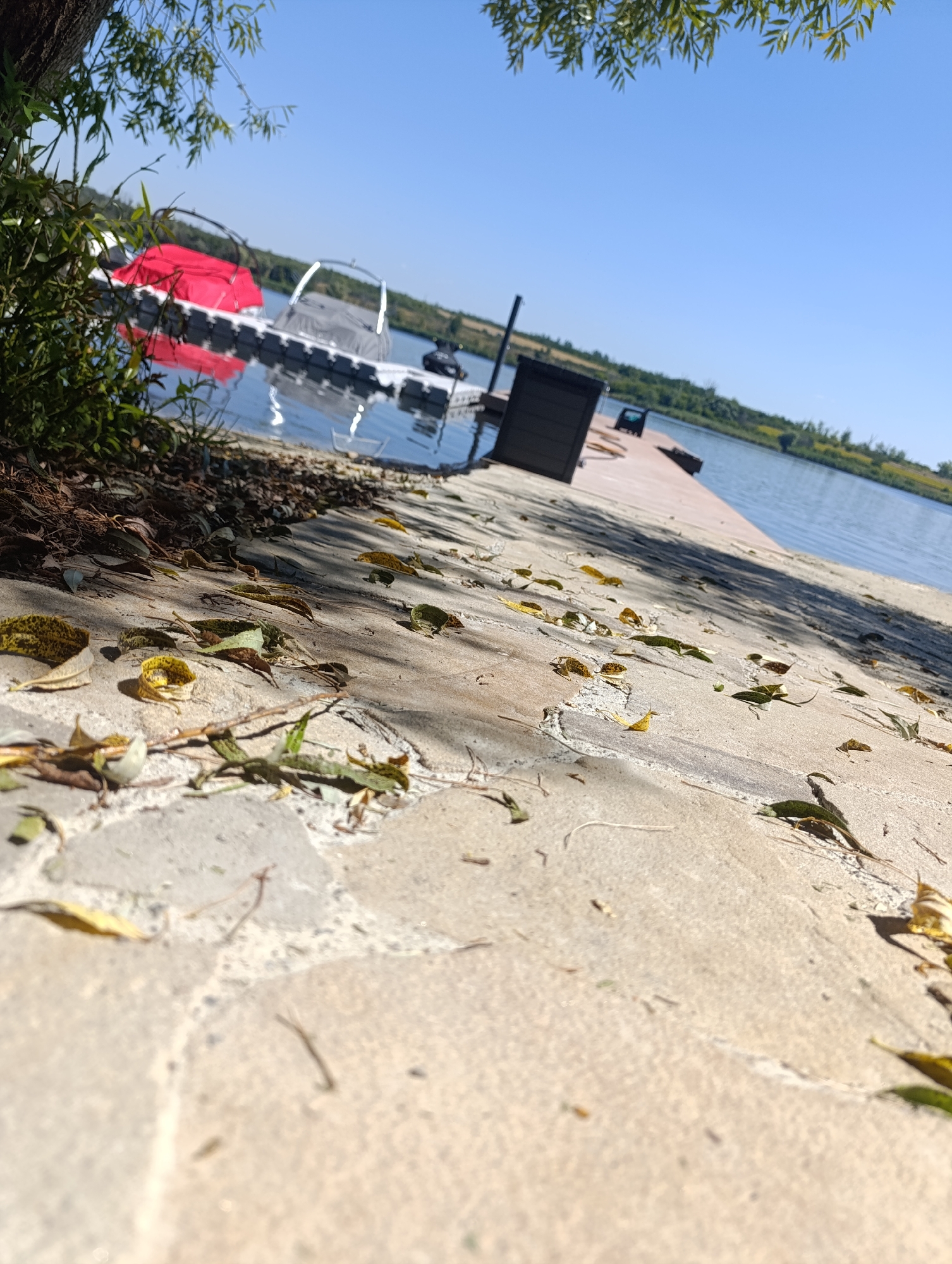 The image depicts a waterfront scene in Bulatselivka, Ukraine, on a sunny day. The foreground is dominated by a stone pathway with scattered fallen leaves, casting shadows. The path leads towards a wooden dock extending into a body of water. On the dock, there is a dark rectangular object, possibly a storage bin or a bench, and further down, another dark object that appears to be a seating area. On the water, adjacent to the dock, are two floating structures. One is a modular pontoon system with a red canopy and a silver-framed canopy, suggesting it might be a small boat dock or watercraft. Another black object is visible further on the pontoon system. The background shows a treeline with greenery and a clear blue sky. The water appears calm, reflecting the sky and the structures on the water. The perspective is low, looking up from the ground towards the dock and the water.