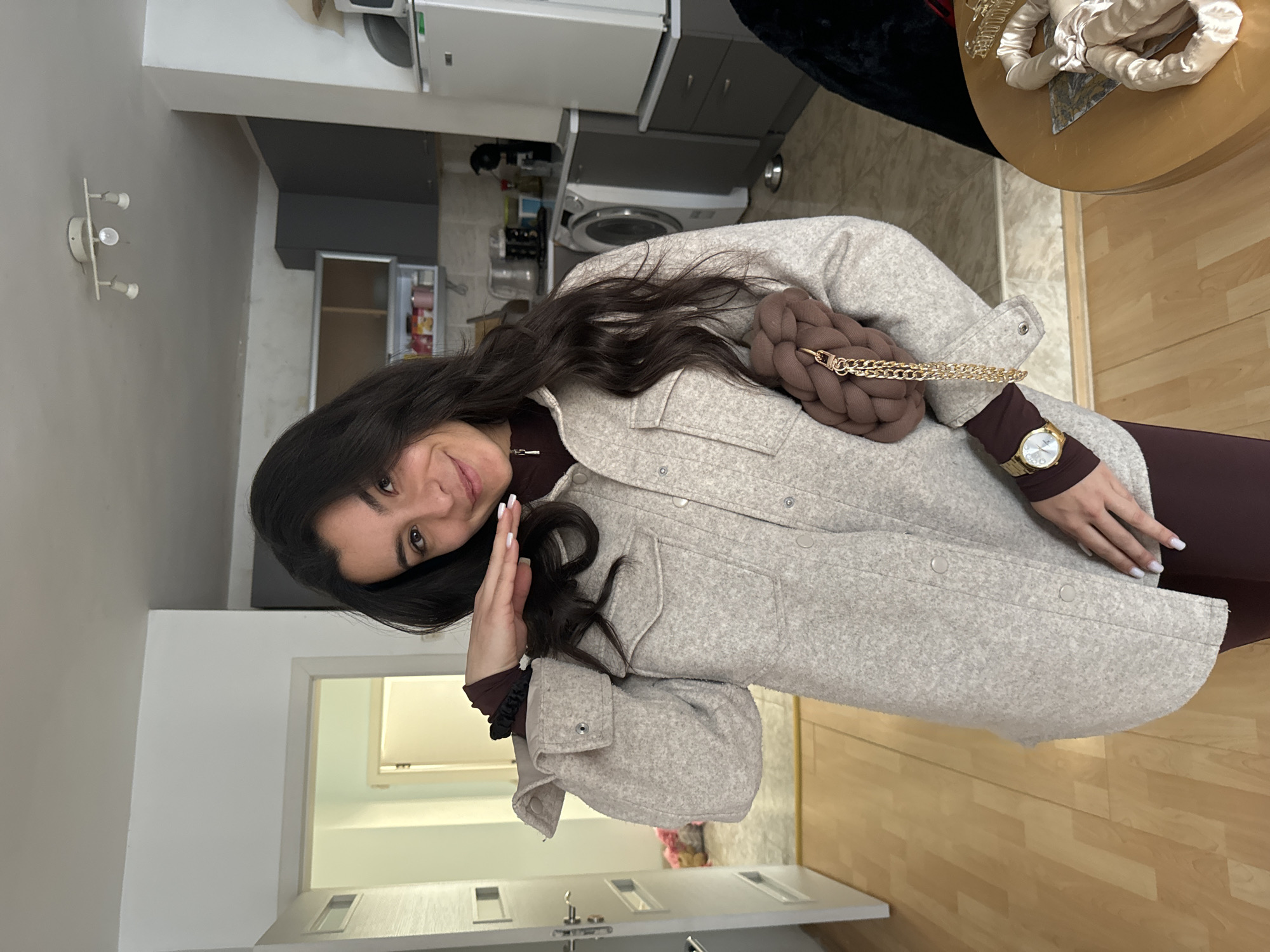 A woman is shown indoors. She has long dark hair, wears a long-sleeved brown dress, a light-colored jacket, and a gold watch. She is carrying a brown braided handbag with a gold chain strap. Her left hand is raised towards her forehead.
The background shows a kitchen with light-colored cabinets, appliances, and a washing machine. A doorway is visible leading to another room. There are no indicators of being in Varna, Bulgaria.