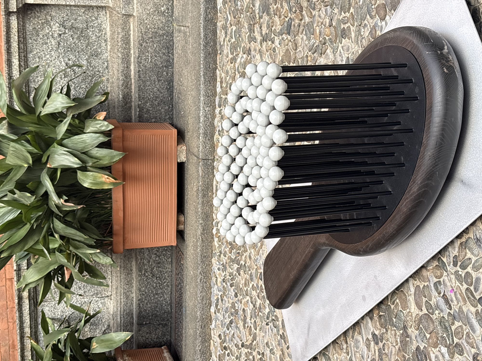 The image displays an outdoor scene featuring a large, abstract decorative object alongside potted plants, set against a textured ground and wall. The main subject, a striking sculptural piece resembling an oversized hairbrush or rake, is positioned on the right. It comprises numerous slender black rods, each topped with a small, white spherical bead, arranged densely in a curved formation. The base of this object is crafted from dark, possibly wooden, material with a smooth, rounded edge, and it rests on a flat, light grey surface.
To the left, several green, leafy plants are arranged in terracotta-colored, rectangular pots. The plants feature broad, vibrant green leaves, some exhibiting slight browning at their tips. Behind the plants, a wall or a series of steps made of speckled grey stone or concrete provides a background element. The ground beneath the decorative object and extending into the lower right of the frame is covered with small, rounded pebbles or cobblestones, creating a highly textured surface.
The scene is brightly lit by natural light, suggesting it is daytime. There are no people visible, and no specific activity is taking place. No discernible text appears in the image. The location context provided for this scene is Milan, Italy.