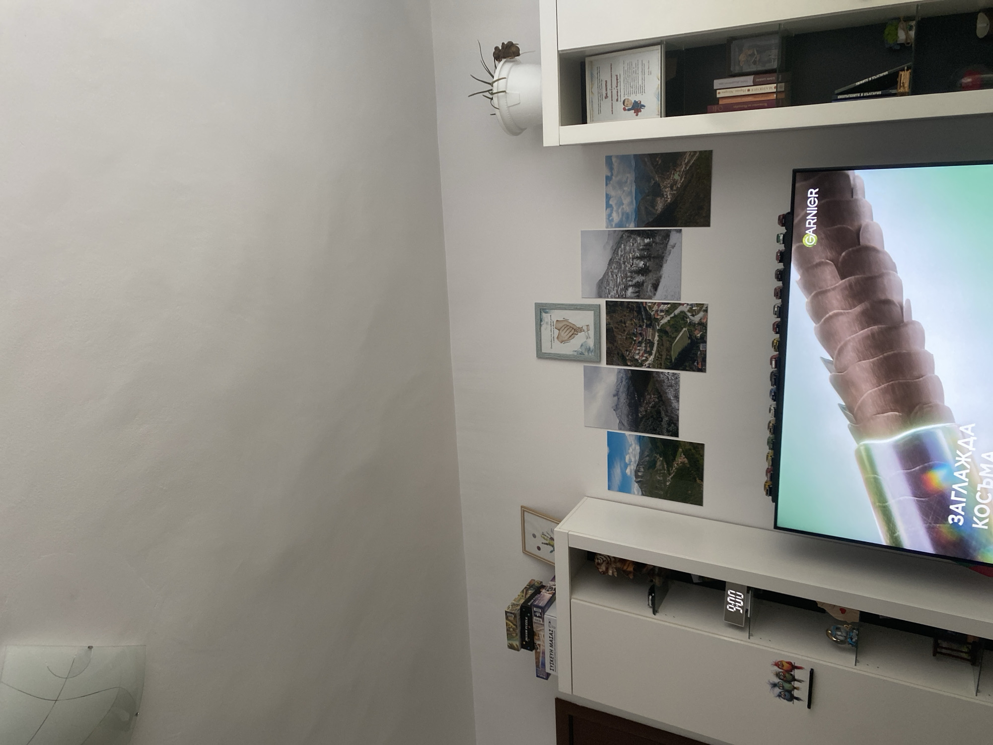This image depicts an indoor scene, likely a living room or entertainment area. The primary focus is on a white cabinet unit on the left, topped with several puzzle boxes and a framed artwork of handprints. Next to it, a white shelf unit displays decorative items including small figurines and a clock displaying "9:00". A large flat-screen television is positioned in front of the cabinets, displaying an advertisement for Garnier, featuring close-up footage of hair strands and text that translates to "SMOOTH HAIR".
Above the television and to the right of the puzzle boxes, a collection of five framed landscape photographs are arranged in a grid. These photos appear to be of mountainous or hilly regions with villages. Further to the right, another white cabinet unit is visible, with a white pot containing a wilting plant on top. This cabinet also holds a framed certificate featuring a cartoon character and text. The shelves below the certificate contain books and other small objects.
The room's walls are painted white, and the ceiling is visible at the top of the image. The overall impression is one of a lived-in, decorated space. There is no direct indication of the city of Plovdiv, Bulgaria, or any specific location within it.