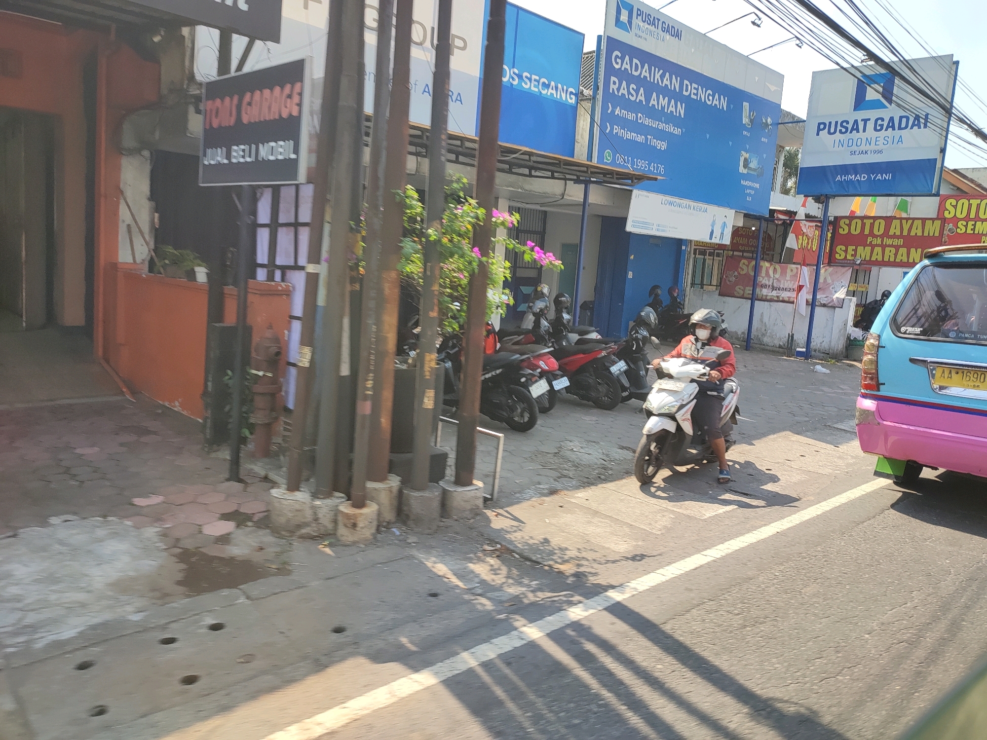 This is a ground-level, slightly elevated outdoor photograph taken in Magelang, Indonesia. The scene depicts a street with businesses on the left and a busy road on the right. The left side features a building with an orange-colored facade and several signs. One sign, in red text on a black background, reads "TOMS GARAGE" and "JUAL BELI MOBIL" (which translates to "Buy and Sell Cars"). Further down the street, a blue building with white signage is visible, with text including "PUSAT GADAI INDONESIA" (which translates to "Indonesia Pawn Center") and "GADAIKAN DENGAN RASA AMAN" (meaning "Pawn with Peace of Mind"). There are multiple motorcycles parked in front of these businesses, and one person is riding a white scooter on the road in the foreground, wearing a red jacket and a helmet. To the far right, the rear of a light blue and pink vehicle is visible, with a yellow license plate that reads "AA 1690 LO". The road surface is asphalt, with a white line dividing the lanes. The lighting suggests it is daytime, with shadows cast by poles and trees.