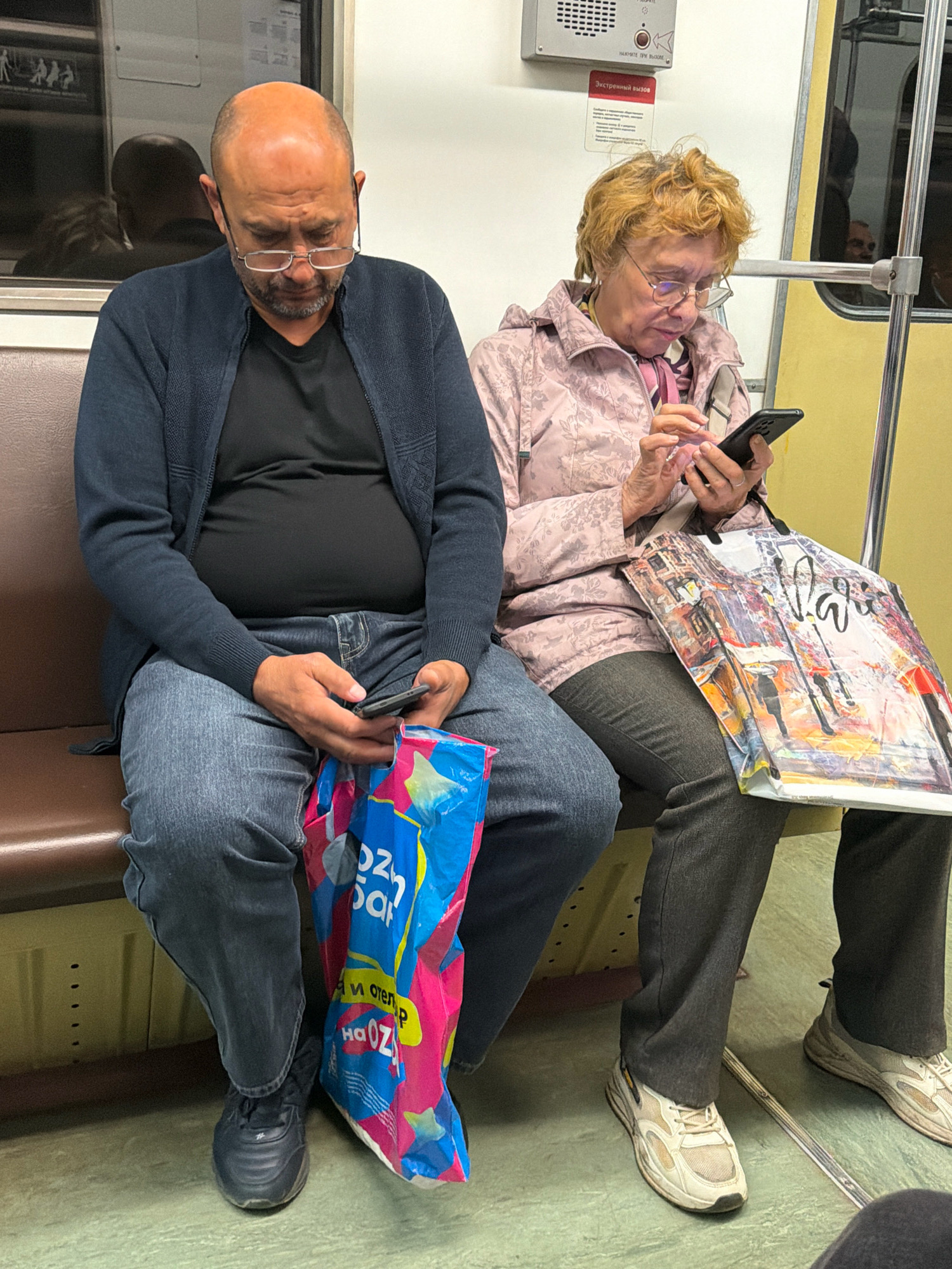 Two individuals, a bald man with glasses and a woman with blonde hair and glasses, are seated next to each other on a Moscow Metro train. The man is wearing a dark blue cardigan over a black t-shirt and blue jeans. He is holding a smartphone in his right hand and a colorful shopping bag in his lap. The woman is wearing a light pink patterned jacket and dark gray pants. She is also holding a smartphone and a large white shopping bag with a Parisian street scene depicted on it. Both individuals appear to be looking at their phones. The environment is a typical subway car, with yellow and cream-colored walls and brown seating.