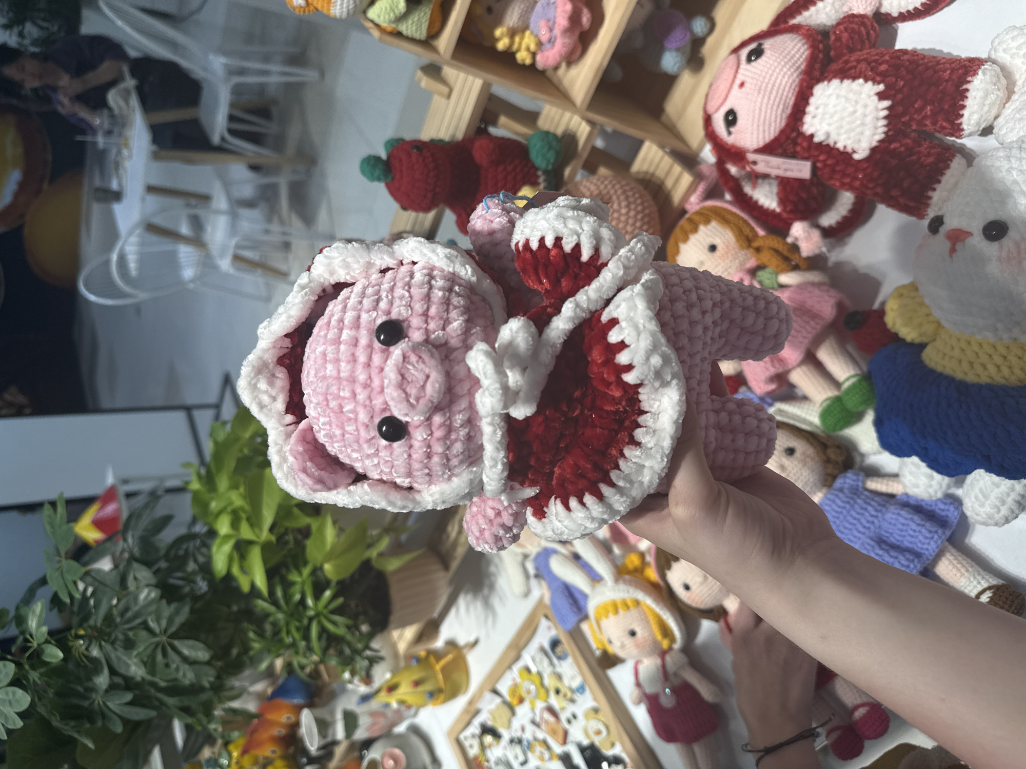 The image shows a person holding a pink crocheted pig toy with a white and red ruffled collar. The background is cluttered with other crocheted toys and plants. A small Vietnamese flag is visible among the plants. There is a wooden display case with more small items, possibly pins or badges. Chairs and tables are visible in the distance. The location is Nha Trang Ward, Vietnam.