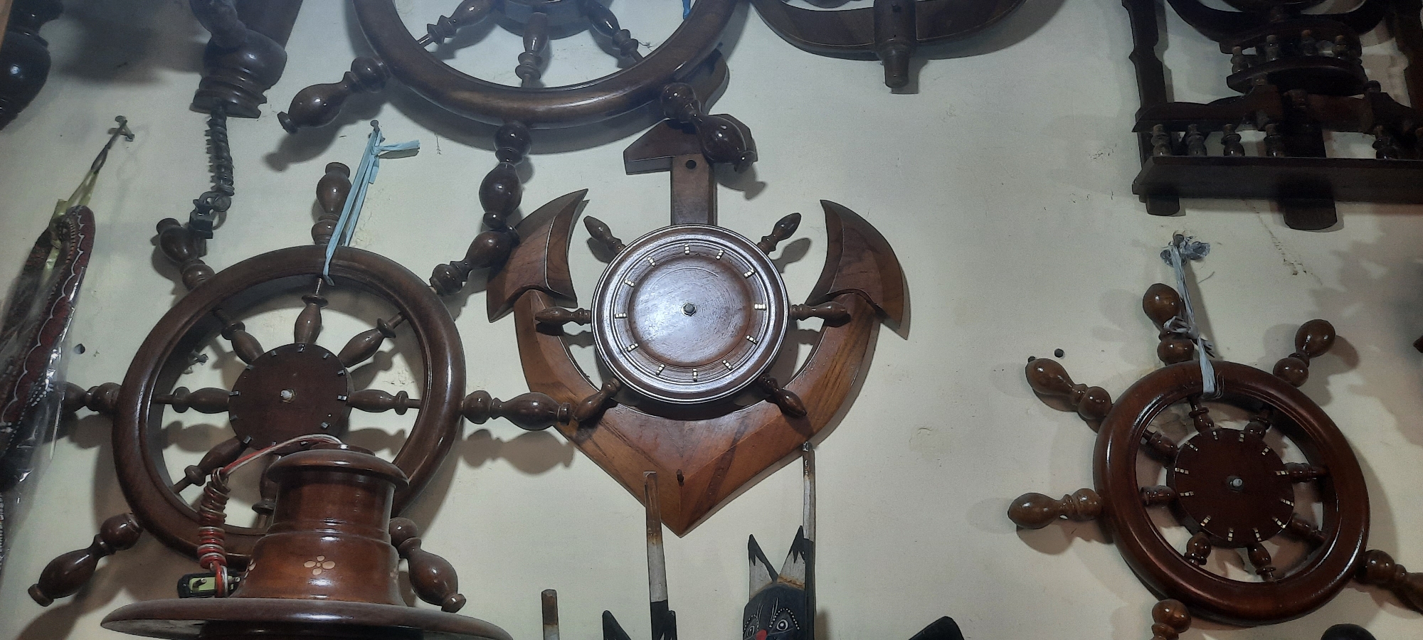 The image depicts a wall adorned with several wooden nautical-themed decorative items, including ship's wheels and an anchor. The items are made of dark polished wood and appear to be handcrafted. The wall itself is a light, neutral color and shows some minor imperfections. In the foreground, a large ship's wheel is partially visible on the left, with a cylindrical wooden object beneath it. In the center, a prominent wooden anchor with a circular detail is mounted on the wall. To the right, another ship's wheel is visible. There are also glimpses of other decorative elements, including what appears to be a wooden stand or furniture piece in the upper right corner, and some decorative items with red and black patterns on the far left. The scene suggests a shop or display area specializing in nautical decor, likely located in Blitar, Indonesia, given the context. No people are visible in the image, and the focus is solely on the arrangement of these decorative objects.