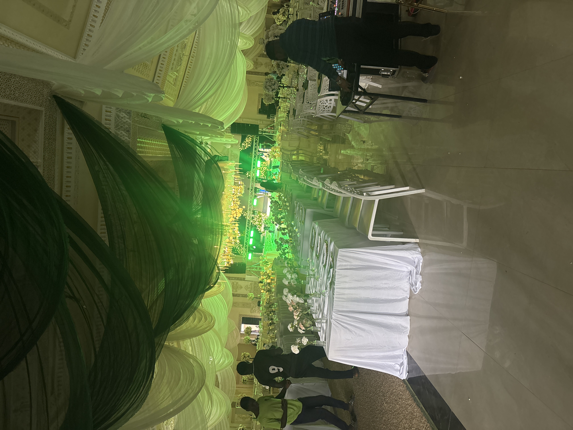 This image depicts a decorated event venue in Jos, Nigeria, likely set up for a wedding reception or a similar formal gathering. The scene is dominated by draped white and green fabrics, creating a luxurious and festive atmosphere. Tables are set with white tablecloths and adorned with floral arrangements in clear vases, suggesting a well-organized event.
Several people are present in the scene. In the foreground, two men are seen walking, one wearing a green t-shirt and the other a dark t-shirt with the number '9' on the back. Further in the background, near a DJ booth with audio equipment, a person is seen operating the controls. The green lighting casts a strong ambient glow throughout the venue, enhancing the visual appeal of the decorations. The floor appears to be a polished, reflective surface, mirroring the lights and decor.
The overall impression is one of preparation or the early stages of an event. The time of day is not explicitly indicated, but the artificial lighting suggests it could be evening or an indoor event. There are no visible weather cues. The setting appears to be a large hall, possibly a banquet hall or convention center, given the scale of the decorations and seating arrangements. The mood is lively and anticipatory, typical of a pre-event atmosphere.