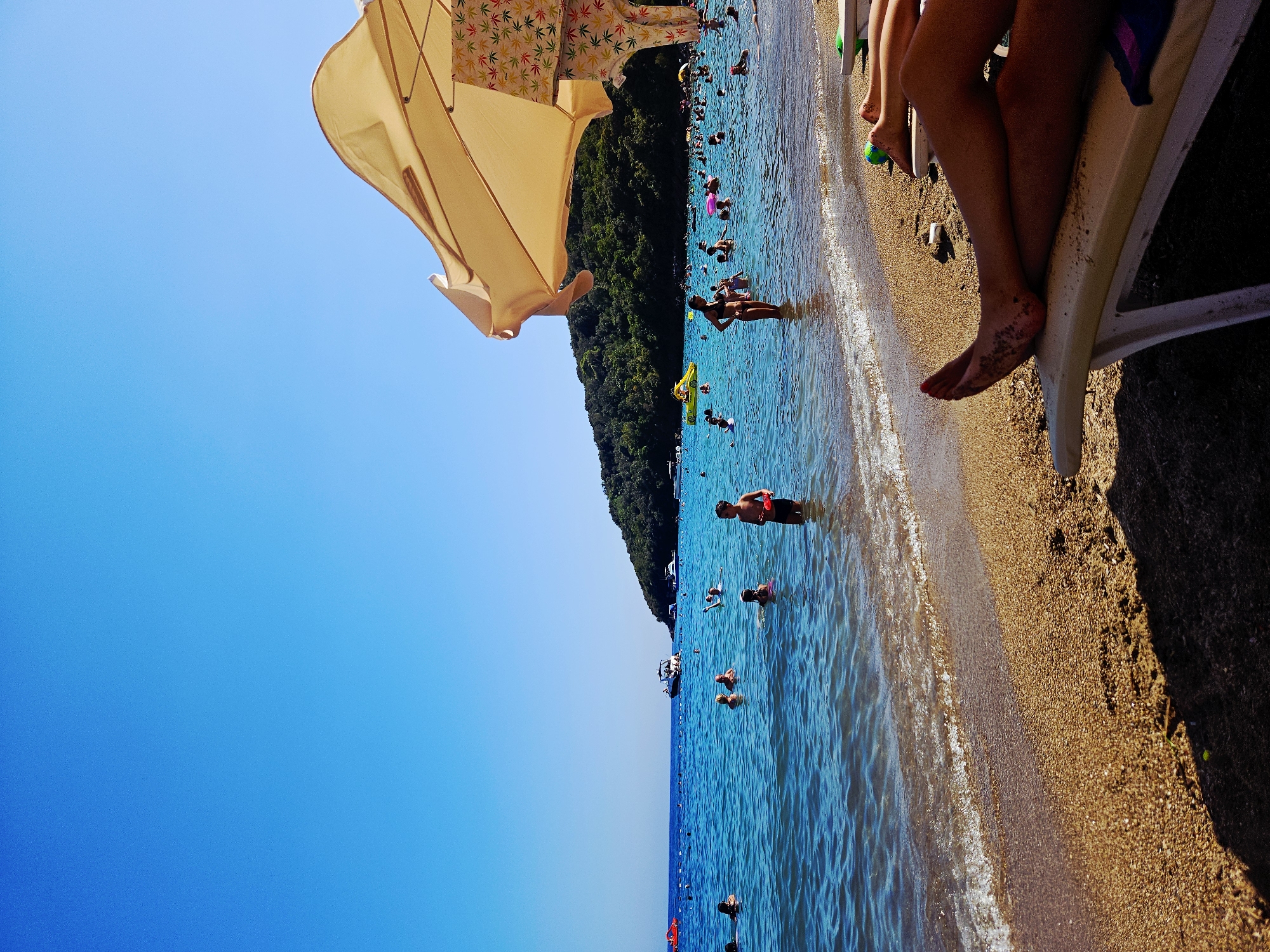 In a crowded beach scene, the azure ocean meets a sandy shore under a clear blue sky. In the foreground, the legs of two individuals are visible, resting on lounge chairs. The water teems with people swimming and wading, some with inflatable rings. A few boats are anchored offshore, and a lush, green hillside forms the backdrop. The image captures a sunny day at the beach, with a vibrant atmosphere and a sense of lively activity.