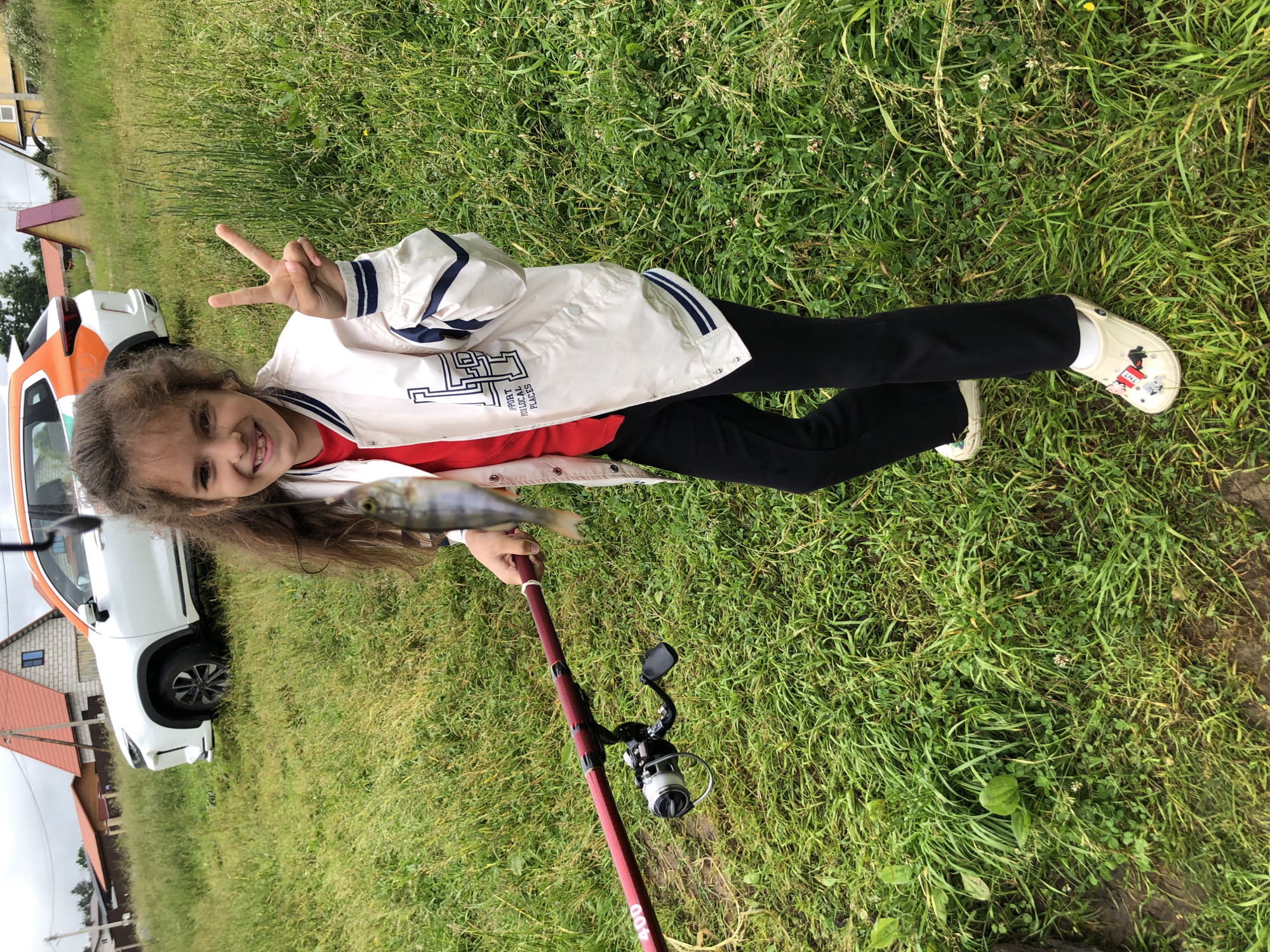 A young girl with dark, wavy hair is holding a fishing rod with a small fish on the line. She is wearing a white bomber jacket over a red shirt and black pants. She is smiling and making a peace sign with her right hand. She is standing in a grassy area with a white SUV and some houses in the background. The location is identified as Bolshiye Polya, Russia. The environment is an outdoor setting with green grass, trees, and buildings.