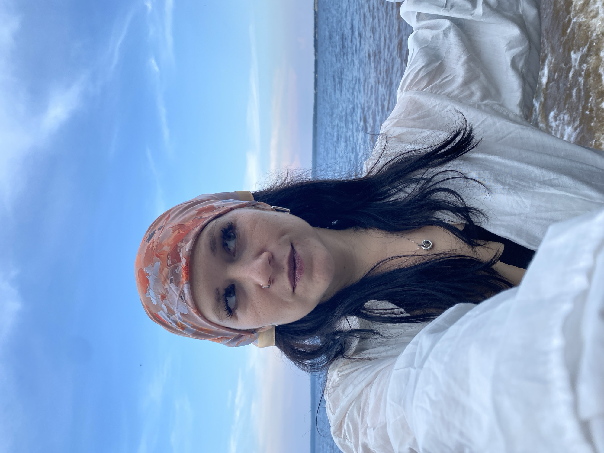 A woman with long dark hair and an orange floral headscarf is standing by a body of water. She is wearing a white, loose-fitting shirt over a black top. The water is choppy and appears to be a river or a large lake. The sky is blue with some wispy clouds. The woman is looking off to the side, away from the camera. The image is a selfie taken from a low angle. This location is the Kama River embankment in Chaikovsky, Russia.